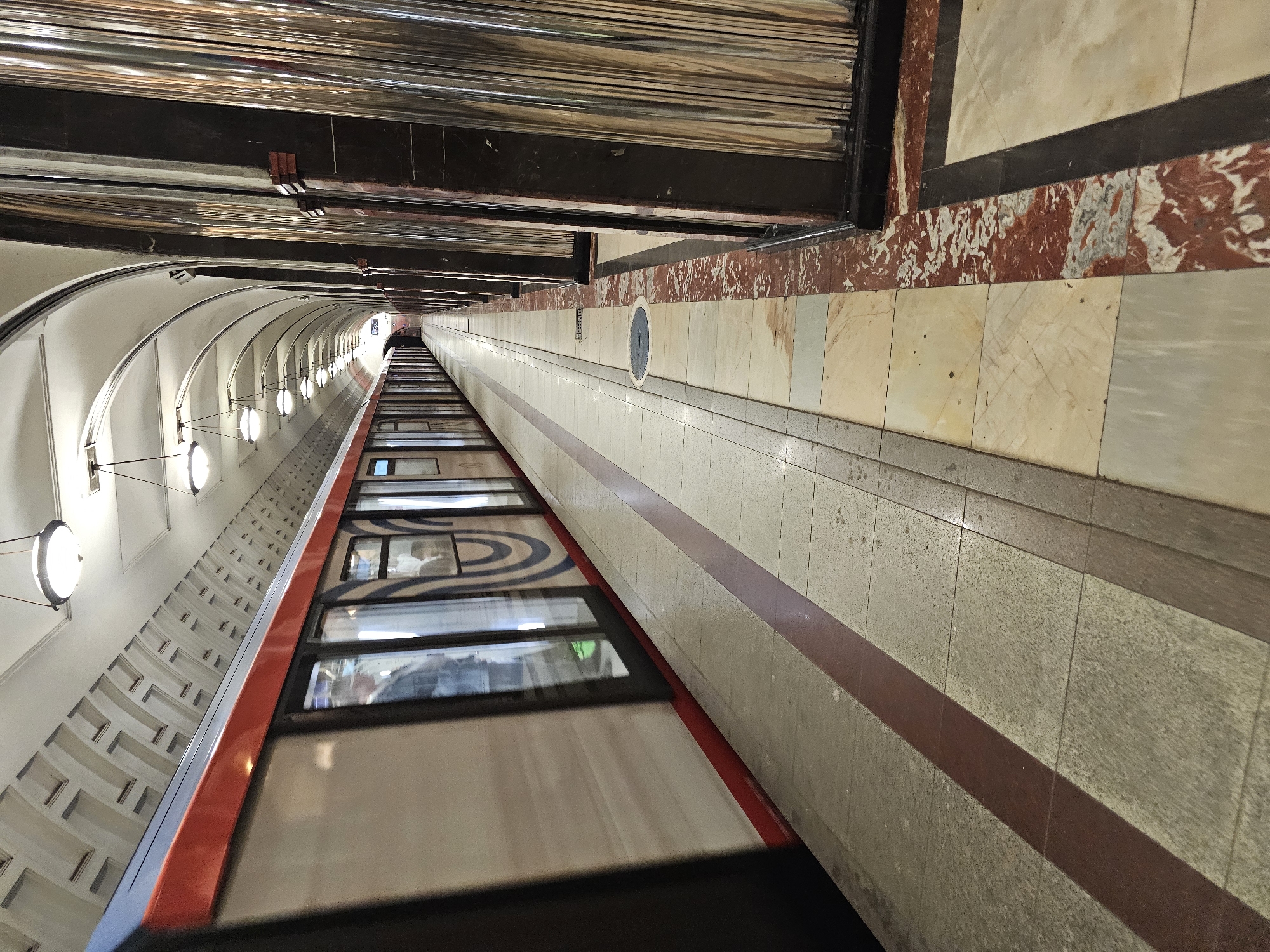 The image shows an angled view of a subway tunnel in Moscow, Russia. The scene is dominated by a subway train with red and grey accents, its windows reflecting the tunnel's interior. Inside the train's windows, blurry figures of passengers can be seen. The tunnel walls are a combination of white, cream, and reddish-brown marble tiles, with a dark horizontal band separating them. Along the left wall, a series of white, circular lights are visible, casting a warm glow. The ceiling features a highly reflective surface, possibly polished metal, with dark horizontal beams. The overall impression is that of a well-lit, modern subway station or tunnel. There are no visible signs of the time of day or weather conditions. The perspective creates a sense of depth, drawing the viewer's eye down the length of the tunnel towards a distant, brightly lit opening.