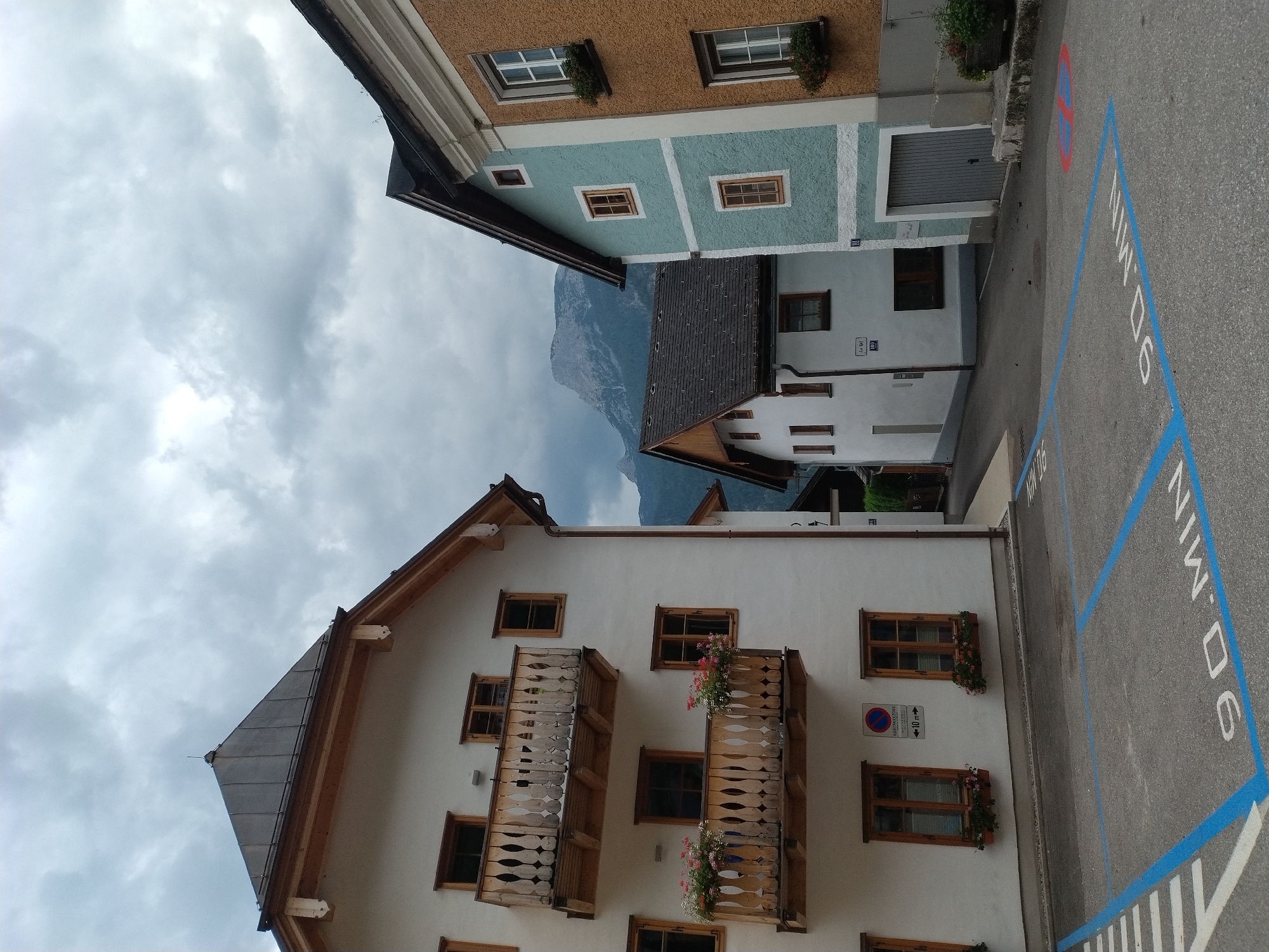 The image shows a street scene in Hallstatt, Austria, with traditional alpine architecture under a cloudy sky. In the foreground, a paved area has parking spaces marked with blue lines and the text "90 MIN" painted on the ground, indicating a 90-minute parking limit. To the left is a white building with a dark sloped roof and wooden balconies adorned with flowers. Two windows are visible on the ground floor of this building. To the right of the white building is a narrow alleyway leading to more buildings. Further to the right, a light blue building stands with a dark sloped roof and several windows. To the far right is an orange-brown building with a similar architectural style and a dark roof. In the background, a mountain range with a snow-capped peak is visible under the overcast sky. The scene is devoid of people.
