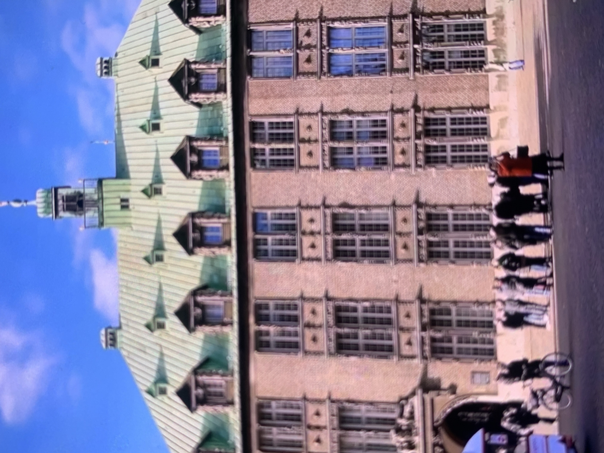 The image depicts a grand, multi-story building with a distinctive green, mansard roof featuring several dormer windows with triangular pediments and decorative carvings. The building's facade is constructed of brick, with elaborate stone surrounds framing numerous windows. The windows themselves are multi-paned and vary in size and arrangement. A prominent tower with a cupola rises from the roofline.
In the foreground, a paved area, likely a plaza or street, is visible. Several people are present, some standing in groups and others walking. A bicycle is parked on the left side of the scene, near a partial view of a blue and white vehicle. One individual in the middle ground is wearing a red jacket and appears to be taking a photograph. The sky above is clear and blue.
While the building's architecture suggests it is an important historical structure, the image does not provide specific visual cues to confirm it is located in Stavropol, Russia.