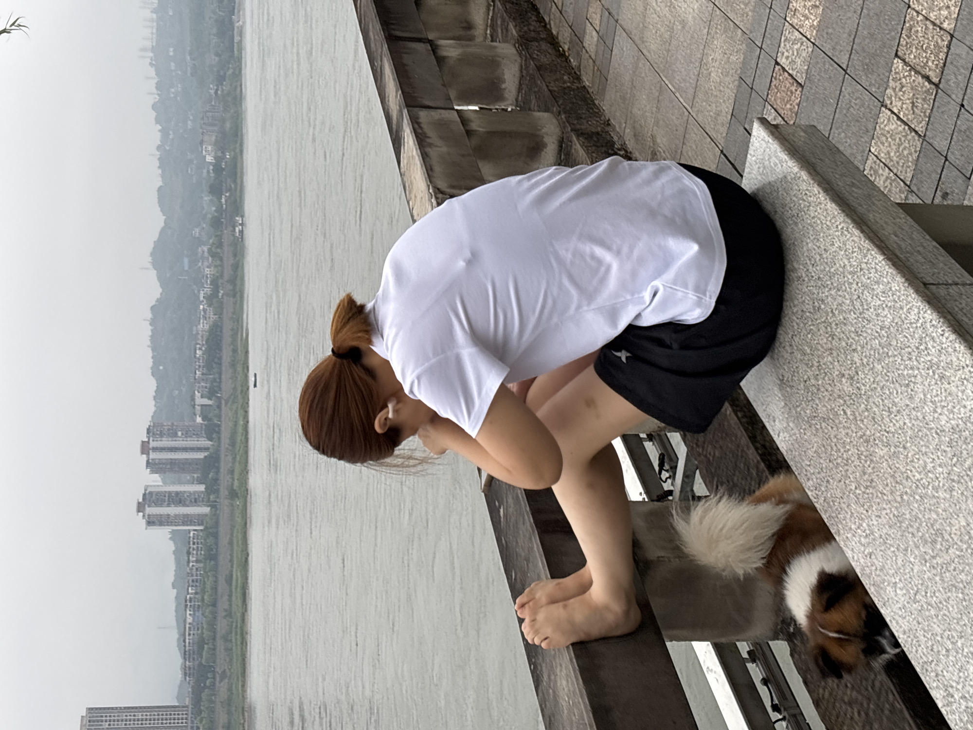 A woman with reddish-brown hair pulled back in a ponytail sits on a granite bench by a river in Jiangjin, China. She is wearing a white short-sleeved polo shirt and black shorts. Her bare feet rest on a wooden railing. A small dog with brown and white fur is partially visible near her feet. The background shows a hazy cityscape with buildings and hills across the river. The sky is overcast. The pavement in the foreground is tiled.