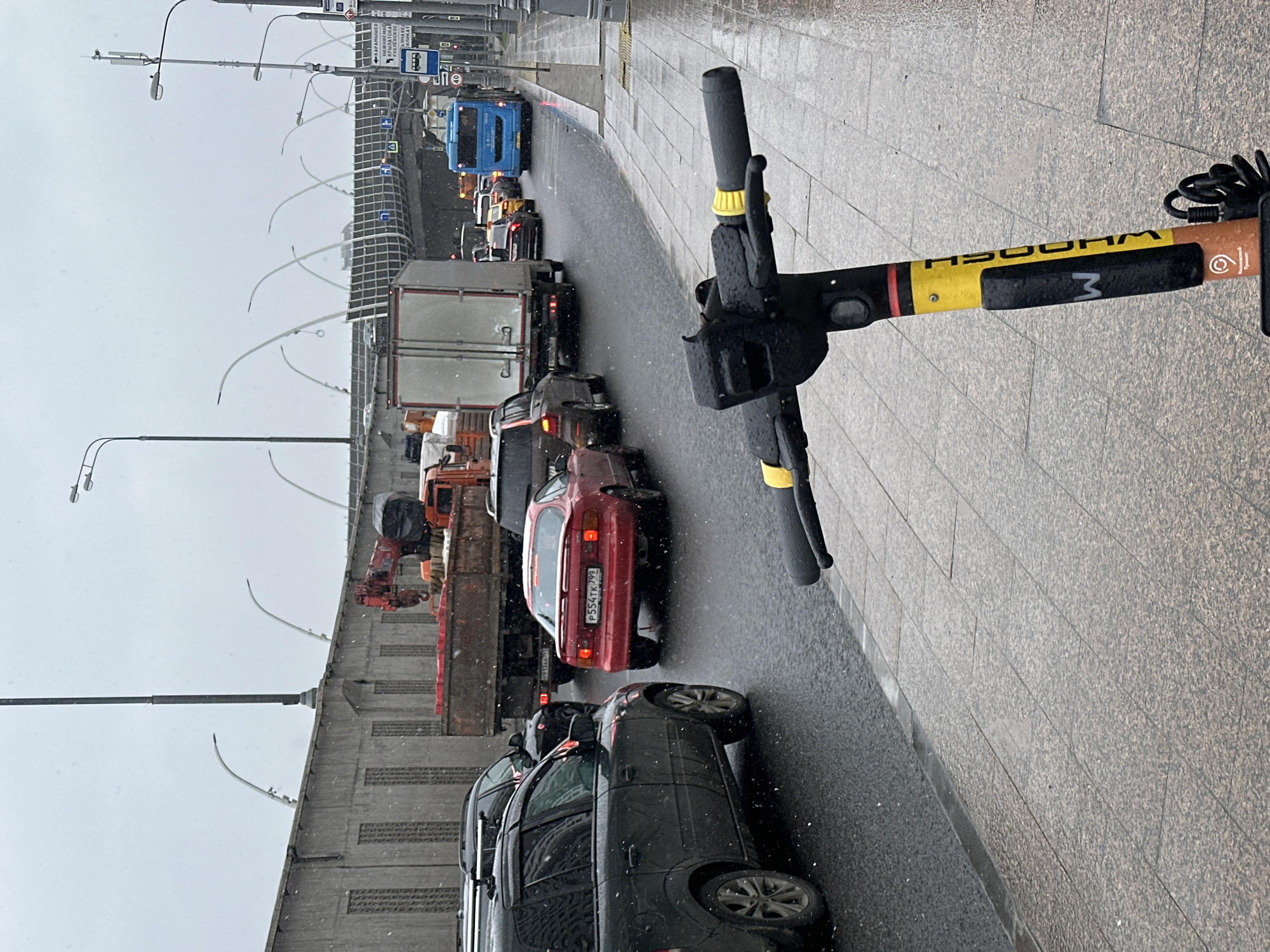 The image shows a traffic jam in Moscow, Russia, viewed from the perspective of someone standing on a sidewalk with an electric scooter. The scooter's handlebars are visible in the foreground. The traffic jam consists of cars, trucks, and a bus, all heading in the same direction on a multi-lane road. A concrete overpass or bridge is visible on the left side of the frame. It is either raining or snowing. The sidewalk is paved with stone tiles. The license plate of a red car in the traffic jam is partially visible, indicating that the image was taken in Russia.