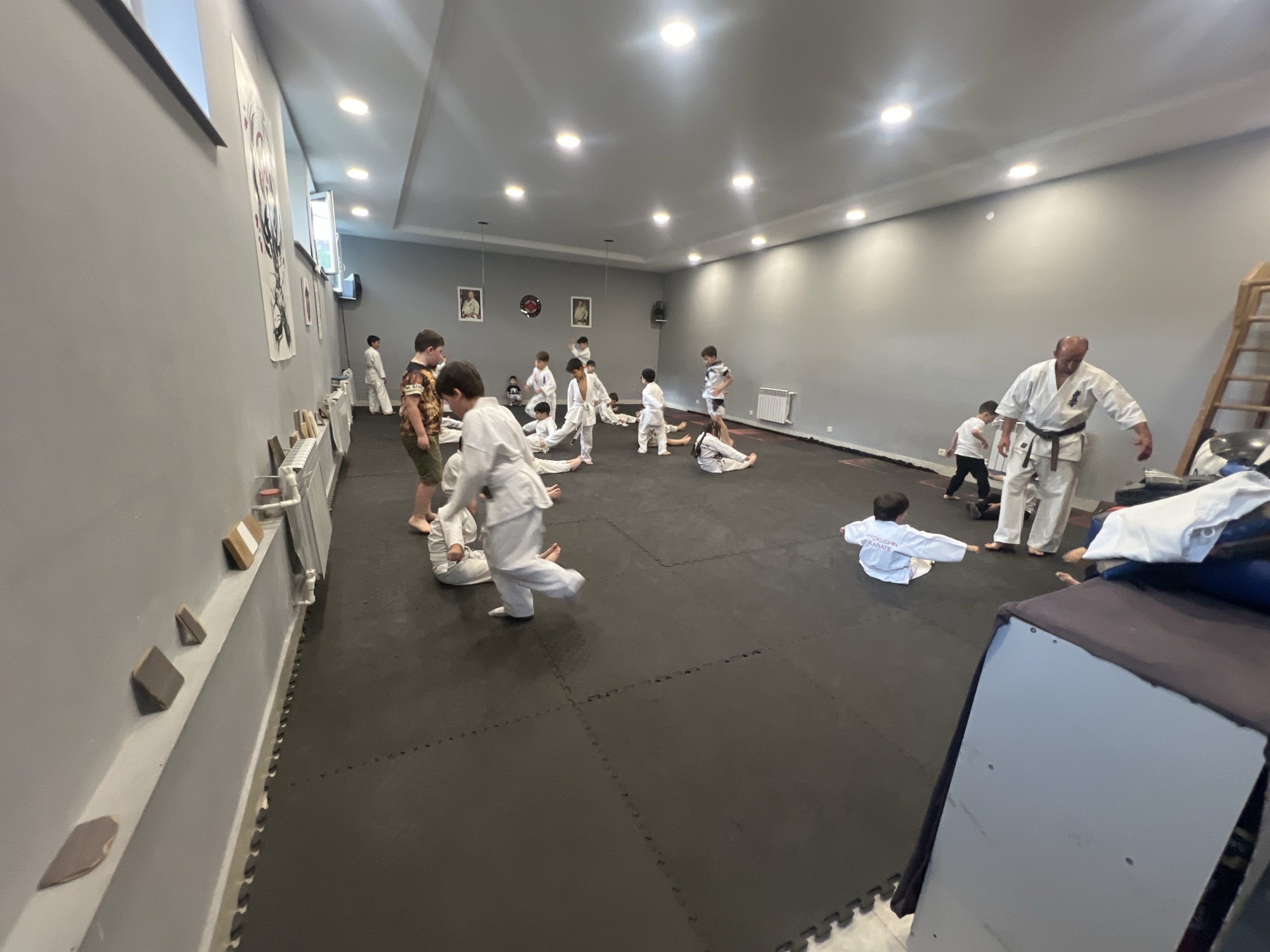 The image depicts an indoor martial arts dojo, likely located in Vanadzor, Armenia, based on available context. The room is spacious with grey walls and a white, paneled ceiling featuring circular light fixtures. A black, padded mat covers the floor, which is characteristic of a dojo.
Several individuals, predominantly children dressed in white karate gi, are engaged in training activities. Some are sitting on the mat, while others are in various stances or in motion. One adult male, also in a gi and wearing a black belt, appears to be an instructor, overseeing the session. The overall atmosphere suggests a lesson or practice is in progress.
On the left wall, there are large framed posters with stylized illustrations, and a radiator is visible along the base of the wall. To the right, a wooden ladder stands against the wall, and a large piece of upholstered equipment, possibly a padded bench or storage unit, is positioned in the foreground. The lighting is bright and even, illuminating the entire space.
