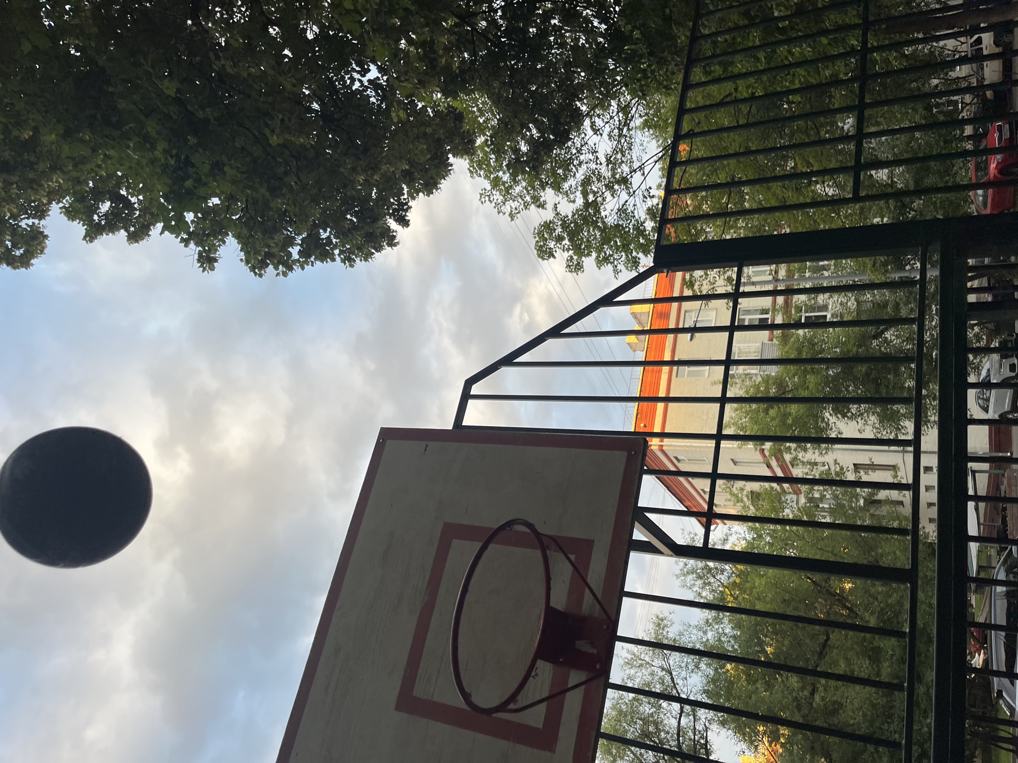 This is a low-angle, outdoor shot looking up at the sky and a basketball hoop in a residential area of Khimki, Russia. The scene is dominated by trees with green foliage at the top and a cloudy sky. A black basketball is suspended in the air on the left side of the frame. In the background, behind a black metal fence, there is a light-colored building with orange trim and several windows. The fence also partially obscures green trees and cars, including a red car. The lighting suggests it is daytime, possibly late afternoon or early evening given the soft light. There is no visible text in the image. The perspective gives a sense of looking up from a basketball court towards the sky, with the urban environment of Khimki visible in the distance.