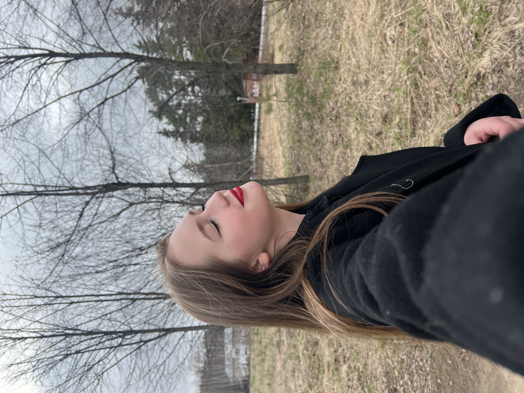 A selfie-style photograph captures a young woman from the chest up, looking upward towards the camera or sky. She has long, light brown hair flowing over her shoulders and is wearing bright red lipstick, with her eyes gently closed, suggesting a serene or contemplative expression. She is dressed in a dark, possibly black, coat or jacket.
The background reveals an outdoor setting dominated by a muted, overcast sky. Numerous bare-branched deciduous trees, typical of late winter or early spring, stand against the grey sky. Further in the distance, some evergreen trees are also visible. The ground is covered with dry, light brown grass interspersed with patches of nascent green, consistent with the seasonal implication of the bare trees. The overall scene conveys a calm, natural environment. No text is visible in the image. The location is contextualized as Chaikovsky, Russia.