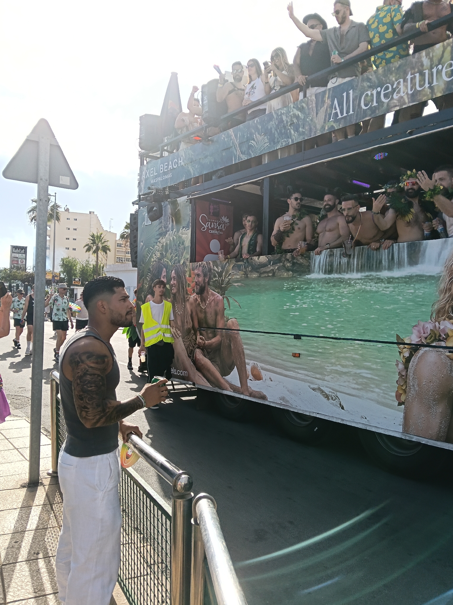 The image depicts a street scene in San Bartolomé de Tirajana, Spain, during a parade or festival. A large, double-decker float with the words "Axel Beach Maspalomas" and "All creatures" is prominent, with people standing on both levels. The float is decorated with a beach-themed graphic. On the ground, a man with tattoos on his arm stands near a metal railing, holding a drink and facing the float. Other people can be seen walking in the background. Buildings and palm trees line the street.