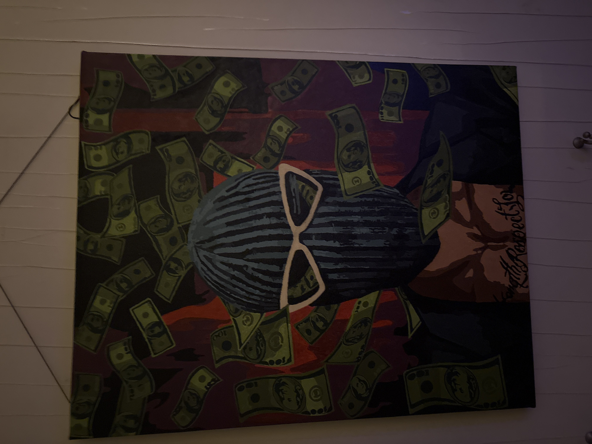 A painting hangs on a light-colored, horizontally paneled wall. The artwork depicts a person's head, obscured by a striped pattern and large, white sunglasses. Falling around the head are numerous green images of US dollar bills. A portion of the person's arm is visible, adorned with a tattoo that includes the words "Loyalty Respect." The background features shades of red and brown. The location appears to be indoors, but there is no further information to suggest the scene is in Ореховка, Russia.