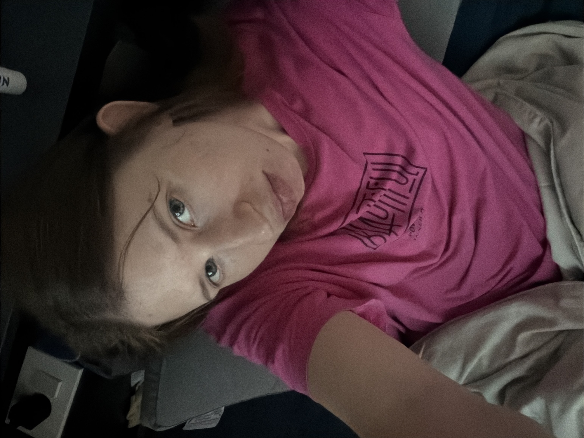 In a dimly lit indoor environment, a young woman with light skin, light brown hair, and blue-grey eyes takes a selfie. She is wearing a vibrant pink t-shirt with a black graphic design on the chest. Her gaze is directed towards the camera, with her head slightly tilted. She is lying down, covered by a beige blanket, and appears to be in a relaxed or reclined position. The background is dark and appears to be a domestic setting, possibly a bedroom or living area, with some indistinct objects visible. The overall lighting is low, casting shadows and creating a moody atmosphere. The image was captured at approximately 11:01 AM. The specific location of Dongducheon-si, South Korea is not discernible from the visual elements of the image.