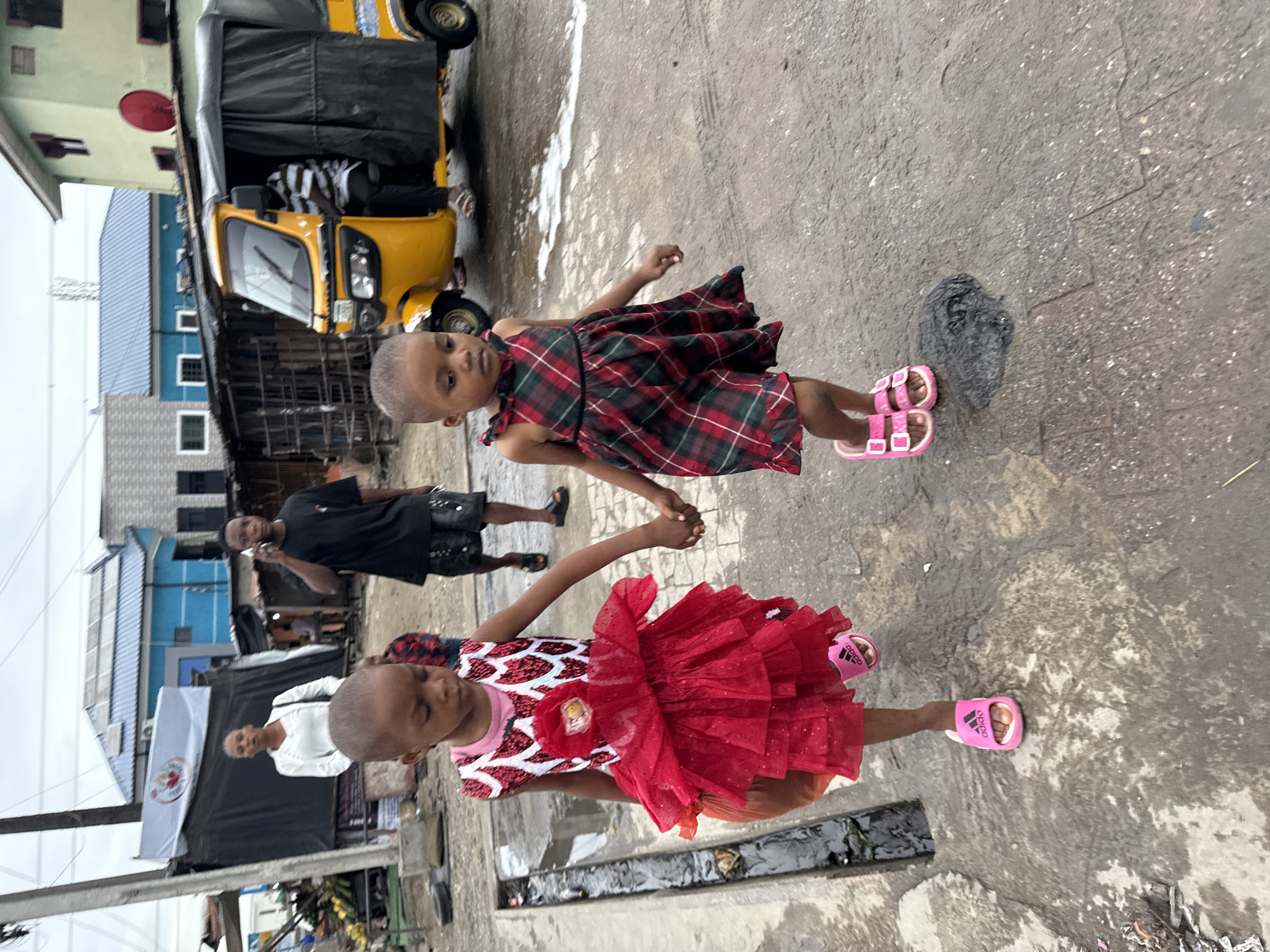 Two young girls are holding hands while standing on a concrete surface in Ado, Nigeria. The girl in the foreground wears a red dress with a white and red patterned top, adorned with a red flower. She has on pink Adidas sandals. The other girl wears a red and green plaid dress and pink sandals.
A yellow auto rickshaw is parked in the background. A man is sitting inside the auto rickshaw. In the middle distance, two men are walking; one is using a cell phone. Various buildings are visible behind the figures. A white flag with a logo is also visible. The street is wet and appears to have a drain.