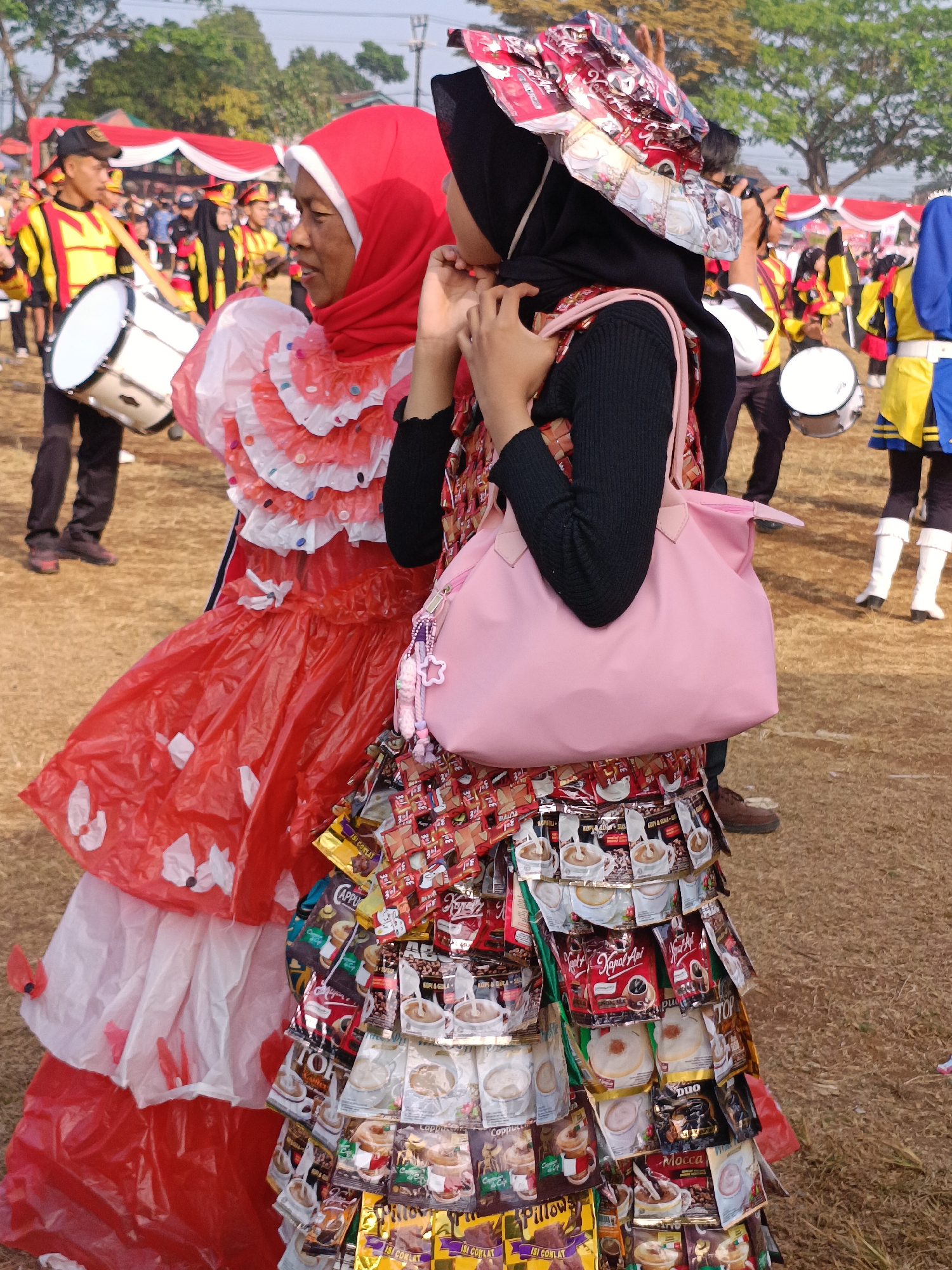 In Cibodas, Indonesia, an outdoor event, likely a parade or festival, is underway. Two women are in the foreground. The woman on the left wears a bright red hijab and a layered red and white dress made of what appears to be plastic sheeting decorated with white floral cutouts. The woman on the right wears a black hijab and a black long-sleeved top. Her skirt is uniquely crafted from numerous individual packets of coffee and chocolate products, creating a textured, layered effect. She carries a light pink handbag adorned with small pink and white beaded charms. Both women are observing something to their left. In the background, a marching band is visible, with several individuals in yellow and red uniforms holding drums. Red and white banners are strung up behind the performers. The ground is covered in dry grass and dirt.