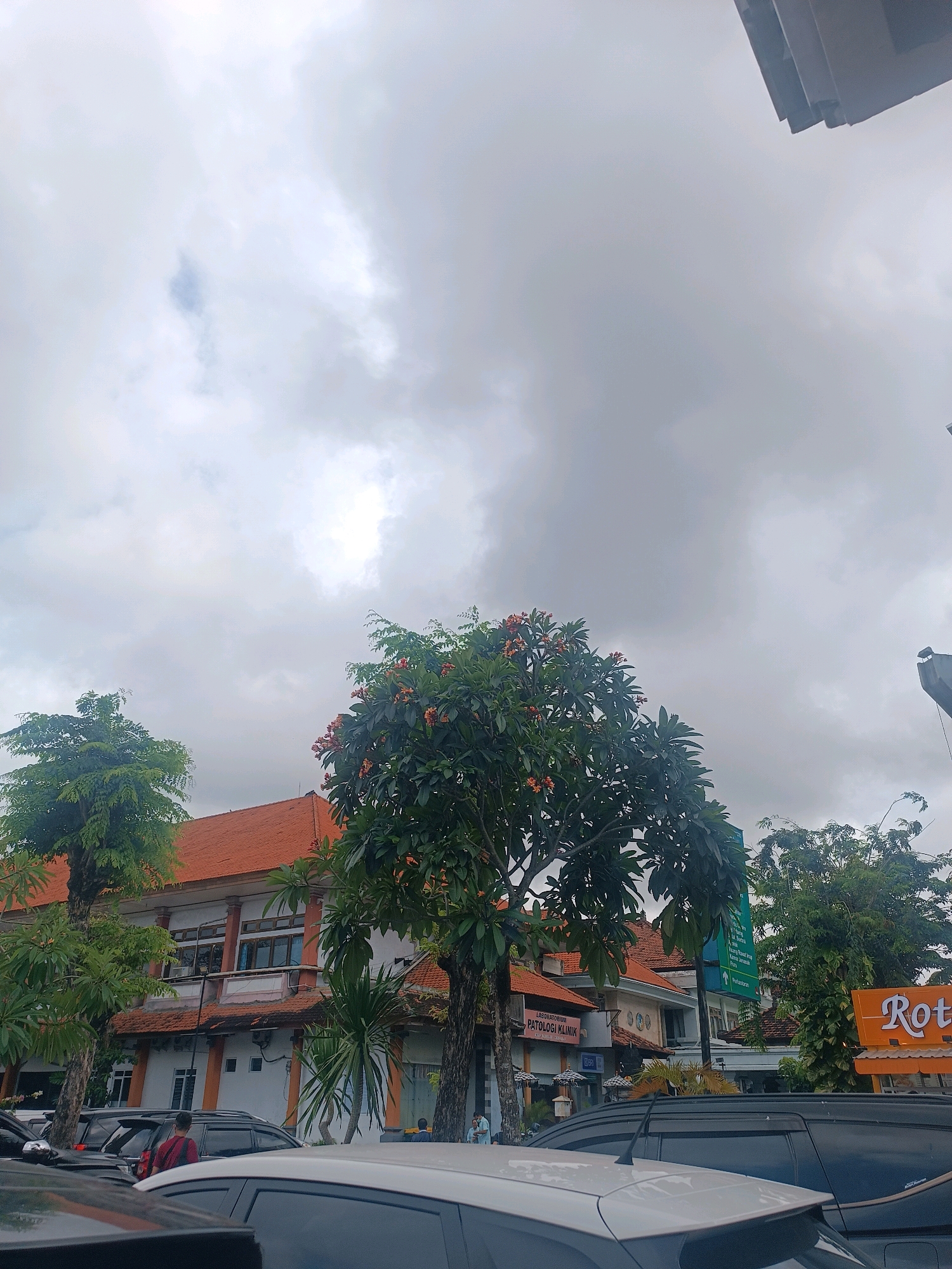 The image depicts a street scene in Denpasar, Indonesia, under a cloudy sky. In the foreground, several vehicles are parked, including a white car in the immediate foreground and dark-colored SUVs behind it. To the left, a building with a terracotta-tiled roof and white walls stands, partially obscured by lush green trees. One tree, in particular, is prominent in the center, bearing small orange flowers. Further to the right, another building is visible, featuring a sign for "Rot." In the middle ground, a street sign in green is mounted on a pole, and to the left of it, a smaller building with a sign reading "LABORATORIUM PATOLOGI KLINIK" is situated. A few individuals are present on the sidewalk, engaged in normal street activities. The overall impression is a typical urban environment in Bali, characterized by its distinctive architecture and abundant greenery, even under overcast conditions.