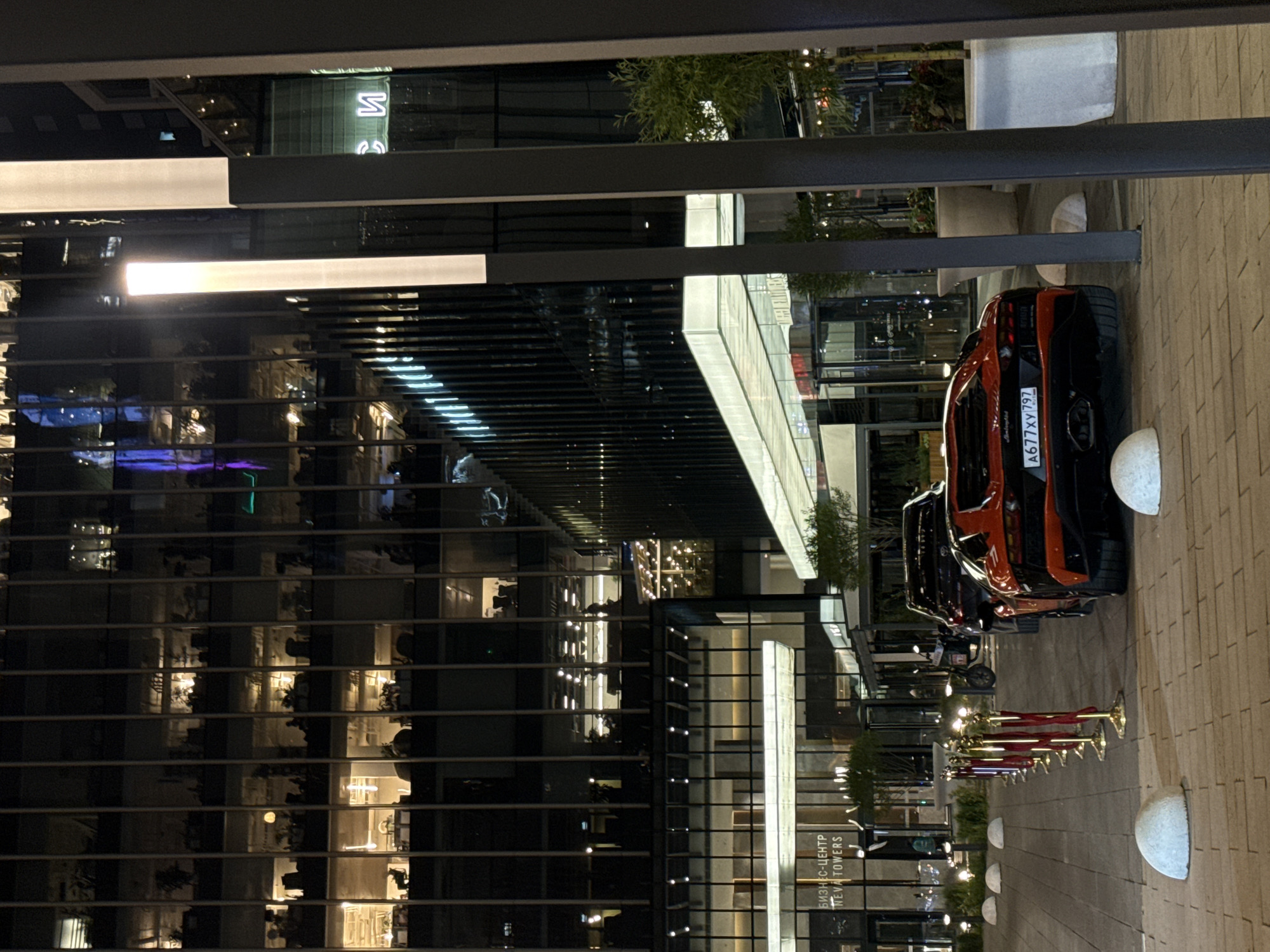 The photograph captures an outdoor night scene in Khimki, Russia, featuring modern architecture and luxury vehicles. A tall building with a reflective glass facade dominates the left side of the image, showcasing a lattice-like pattern of dark window frames. The reflection reveals internal illumination, suggesting office spaces or residences within.
On the right side, a row of luxury cars, including what appears to be an orange Lamborghini, are parked along a paved walkway. The license plate of the Lamborghini reads "A677XY797". Ropes, likely decorative barriers, line the pathway. The scene is lit by both the artificial lights of the building and what appear to be streetlights along the walkway. A sign in Russian, "БИЗНЕС-ЦЕНТР NEVA TOWERS", is visible at the base of the building. No people are prominently featured in the composition.