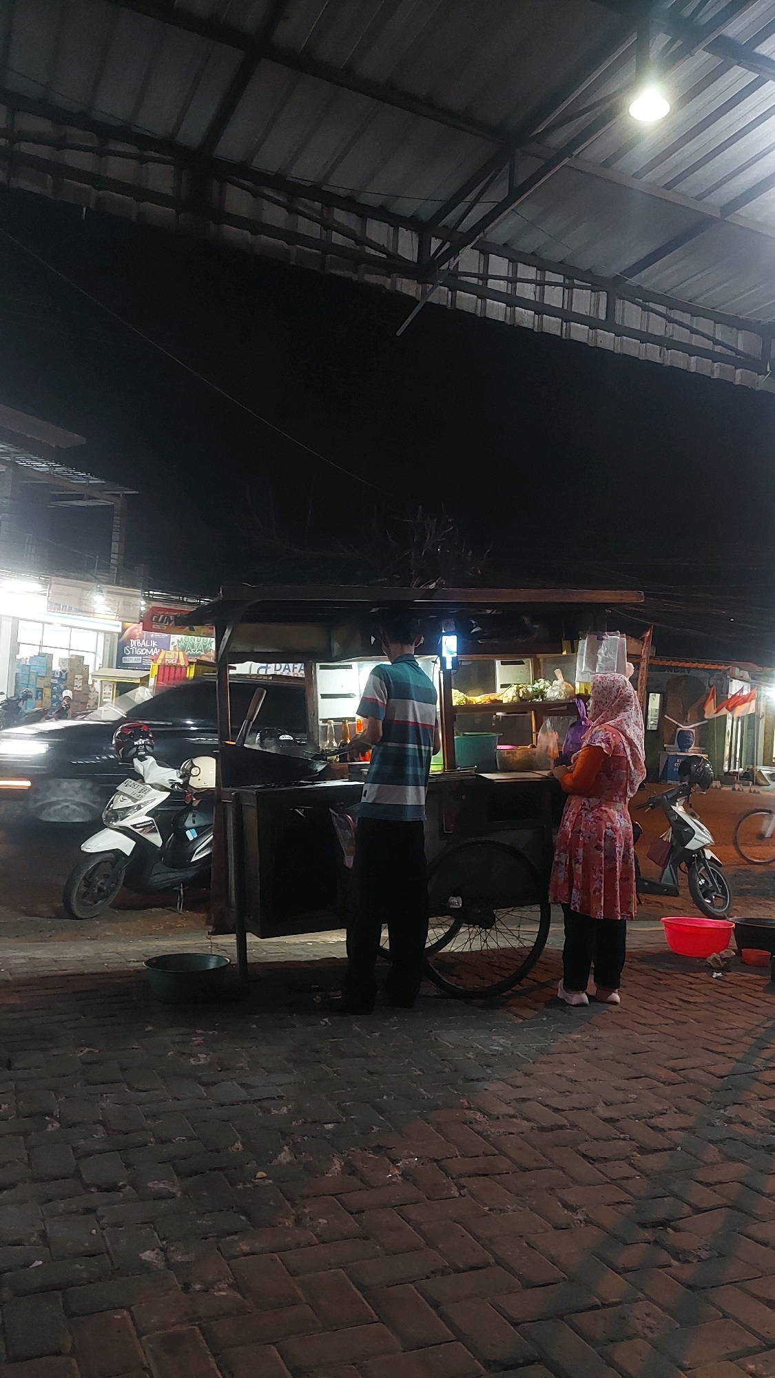 A nighttime outdoor scene in Banyudono, Indonesia, features a food cart at a street-side market. A man in a striped t-shirt stands behind the cart, appearing to prepare food. A woman wearing a headscarf and a floral dress stands to his right, holding a plastic bag. The food cart is equipped with a stove, various containers, and shelving. A black SUV is parked to the left of the cart, and a white scooter is parked in front of it. The ground is paved with dark bricks, and a large red bucket is visible on the right side of the frame. Overhead, a corrugated metal roof supported by metal beams is illuminated by a single light fixture. The overall ambiance suggests a bustling local market at night.