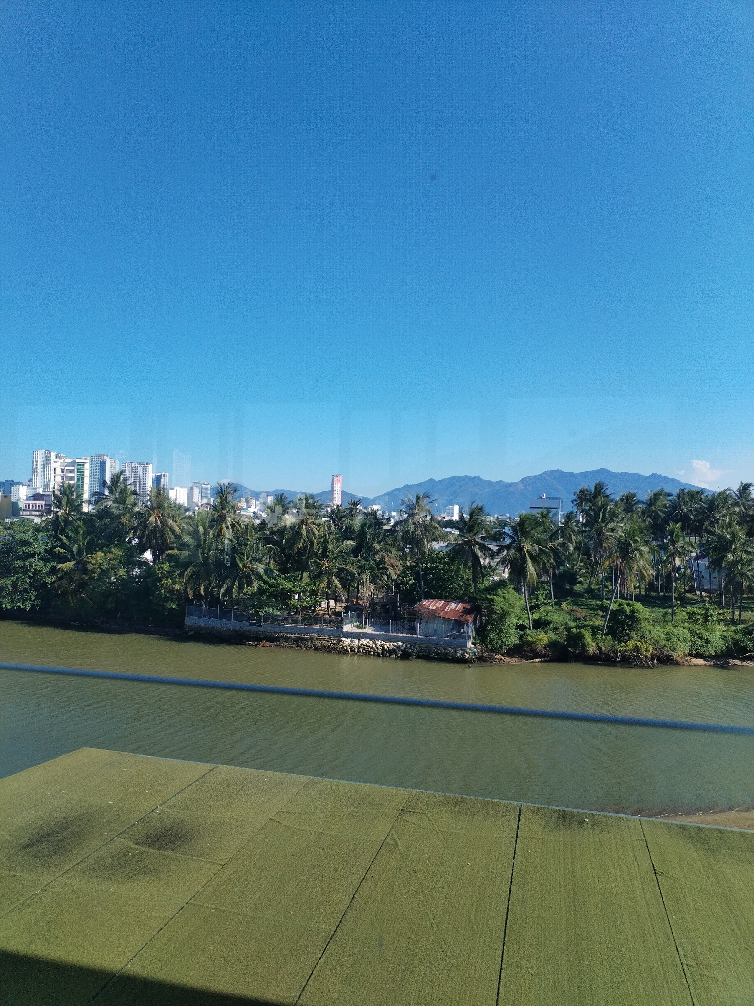 This is a landscape photograph taken on a clear, sunny day in Bac Nha Trang Ward, Vietnam. The image captures a wide view of a river in the foreground, with a lush green bank lined with palm trees and other vegetation. In the distance, a city skyline with numerous high-rise buildings is visible, backed by a mountain range. A small, rustic building with a rusty roof is situated on the riverbank among the trees. The sky is a vibrant blue, with no clouds in sight. The foreground of the image features a large, flat, green area, possibly a rooftop or a recreational surface, divided into sections by dark lines.