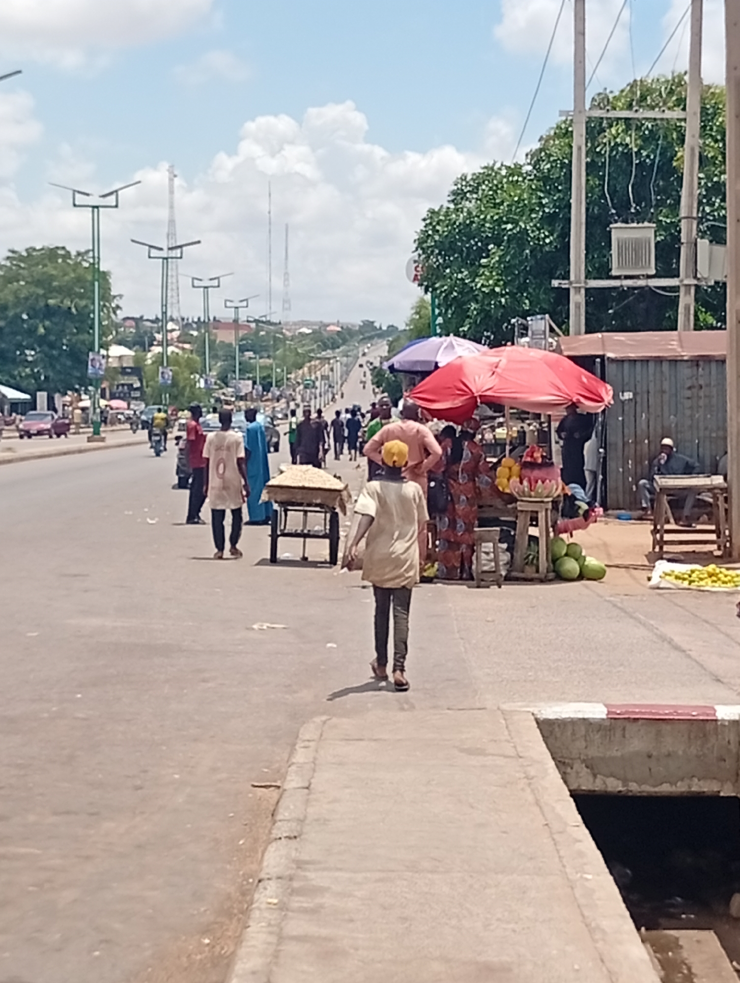 The image depicts a street scene in Tumfure, Nigeria, likely on a market day. A busy road runs through the center, with several lanes of traffic visible in the distance, including cars and motorcycles. Pedestrians are walking on the side of the road and on the pavement. A market stall is set up under a red umbrella, selling fruits and other goods. There are several people gathered around the stall, some appearing to be vendors and others customers. In the background, more market stalls and shops can be seen lining the street, along with tall utility poles and trees. The sky is partly cloudy, indicating a bright day. The overall impression is one of a vibrant and active urban environment.