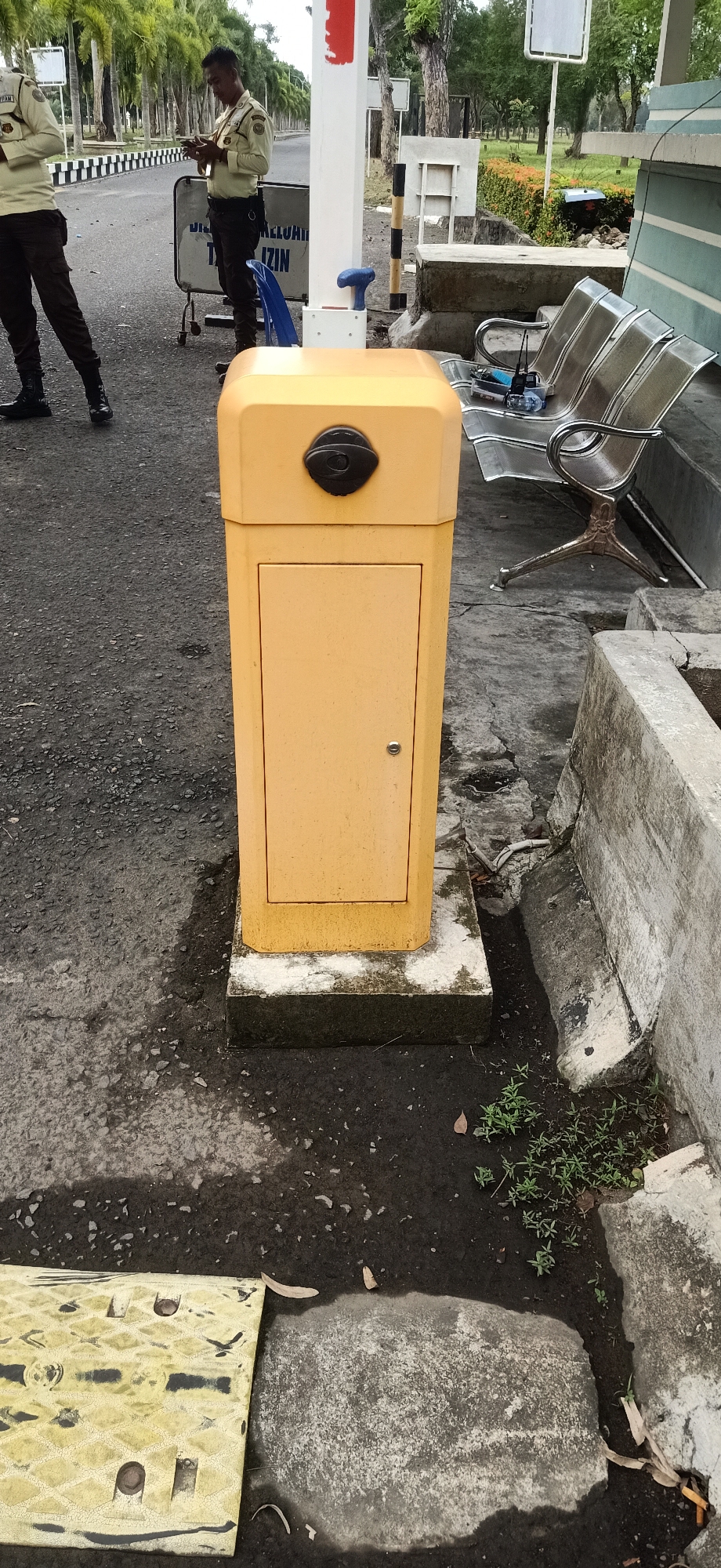 At the entrance to a facility in Tambontunong, Indonesia, a yellow automated barrier gate stands on a concrete pedestal. Two security guards in light-colored uniforms are positioned near a barrier with black and white stripes. One guard, facing away from the camera, is holding a mobile phone. The ground is paved with asphalt, and a yellow speed bump is visible in the foreground. In the background, trees line the road, and a row of metallic benches sits next to a concrete structure.