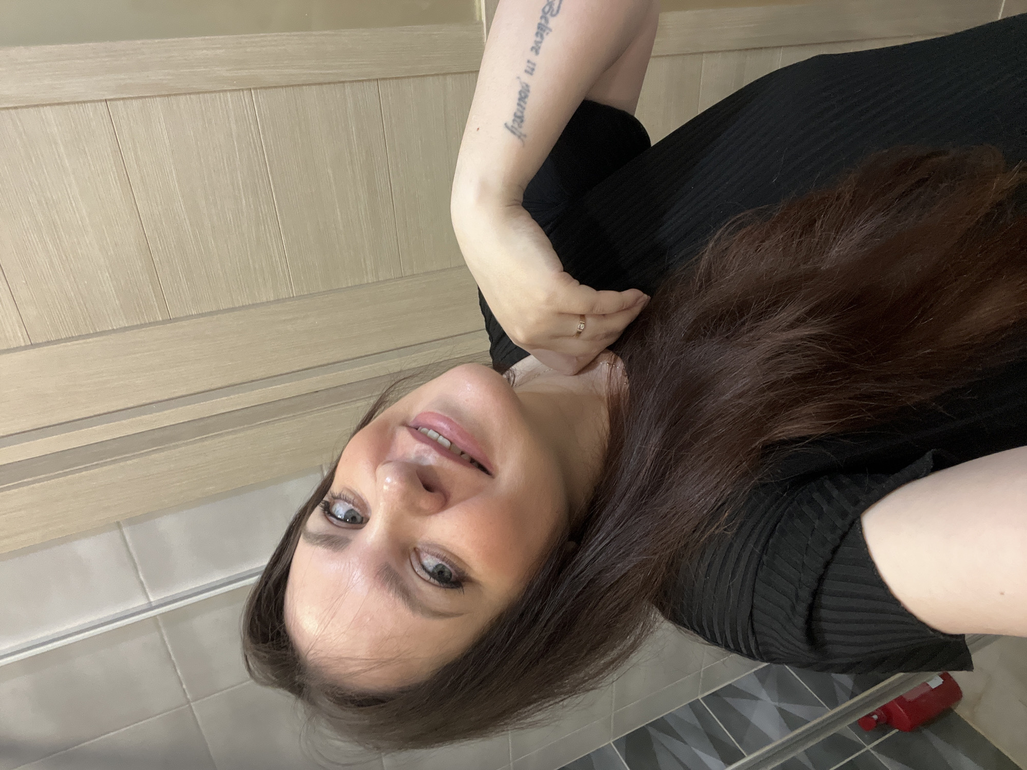 A woman with brown hair and fair skin is seen in a tilted close-up shot. She is wearing a black top, and a tattoo that reads "Believe in yourself" is visible on her left arm. She has a ring on her finger. The background shows a tiled wall and a wooden-paneled door. A red bottle is visible in the lower right corner. There's nothing in the image to indicate that the location is Noviy, Russia.