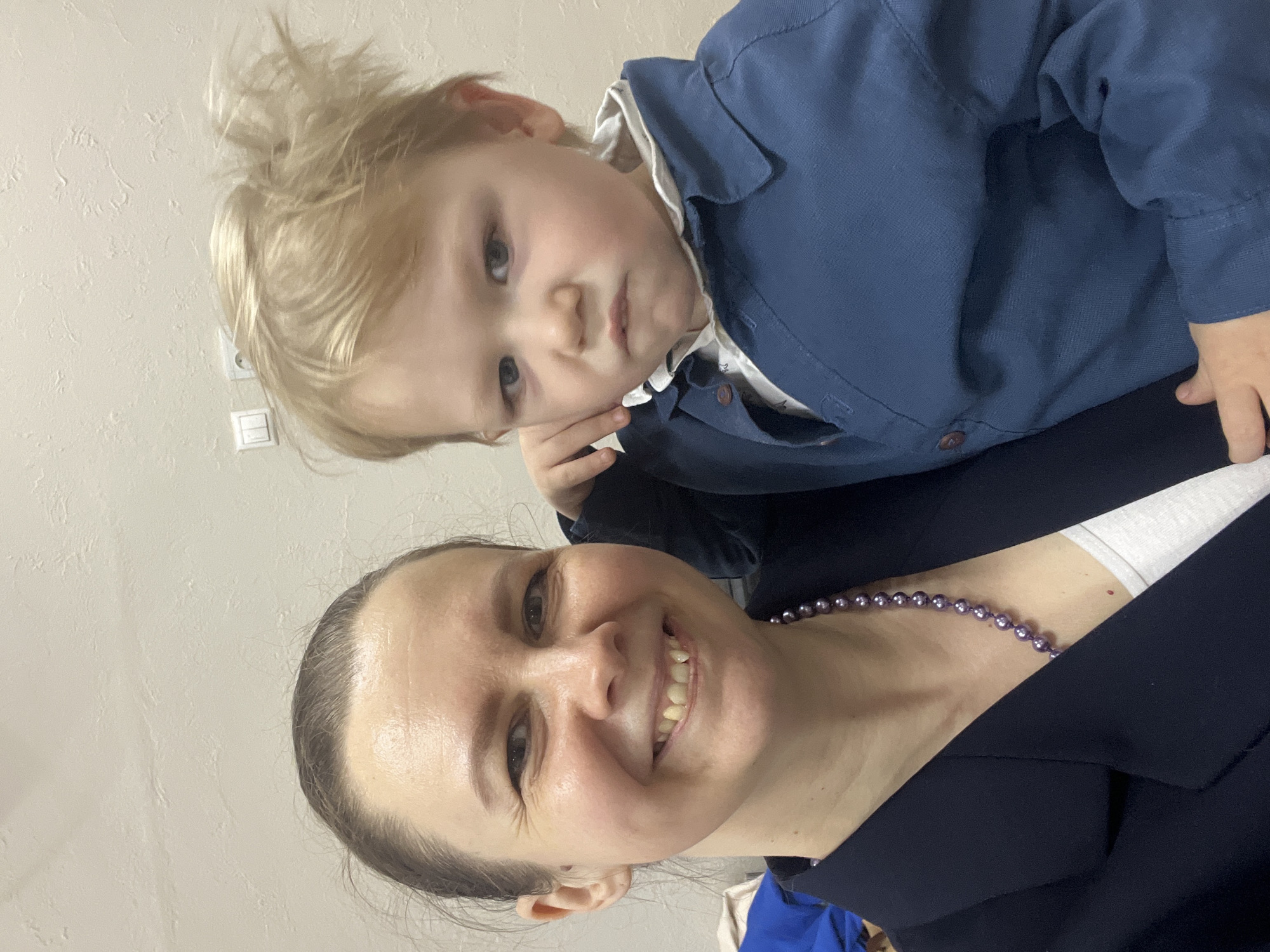 A woman with light skin and dark hair pulled back is smiling broadly at the camera. She is wearing a dark blue blazer and a purple beaded necklace. On top of her shoulder, a young child with blonde hair and fair skin is looking at the camera. The child is wearing a blue button-down shirt with a white collar. A light-colored wall is in the background. An electrical outlet is partially visible on the upper left. The photo was taken inside a building located in the city of Chaikovsky, Russia.
