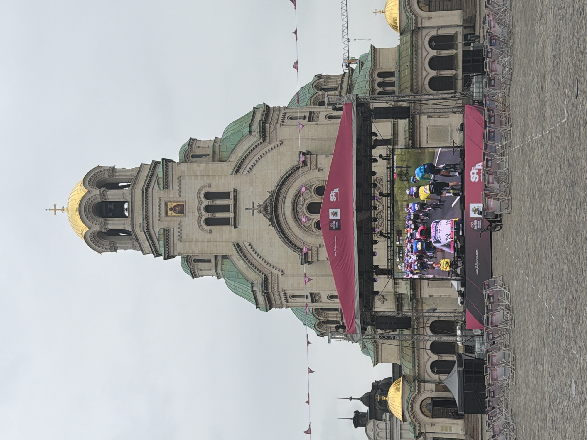 The image shows the Alexander Nevsky Cathedral in Sofia, Bulgaria. A large screen stands in front of the cathedral, displaying a cycling race. Banners and pennants are hung around the screen. Rows of chairs are arranged in front of the screen, suggesting an audience. The sky is overcast. The ground is paved with stone blocks.
