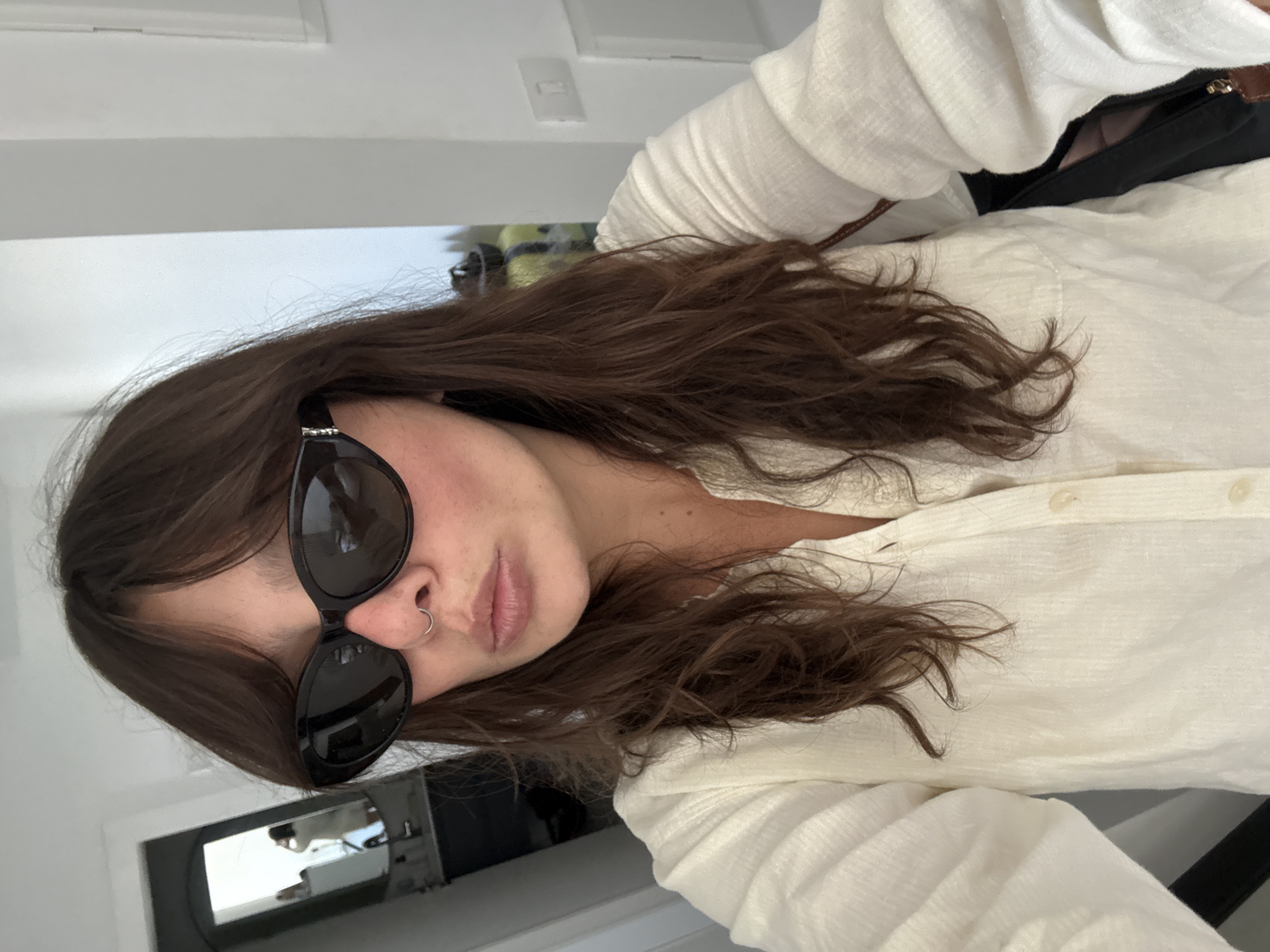 A woman is taking a selfie in what appears to be a hotel room. She is wearing sunglasses, a nose ring, and a cream-colored linen shirt. Her hair is brown and wavy. In the background, a mirror reflects another person, possibly taking a picture. The room is brightly lit with white walls and a doorway leading to what looks like a bathroom. The overall setting suggests a casual, possibly vacation-related environment. It is impossible to determine the specific location within Tel Aviv based on the provided image.
