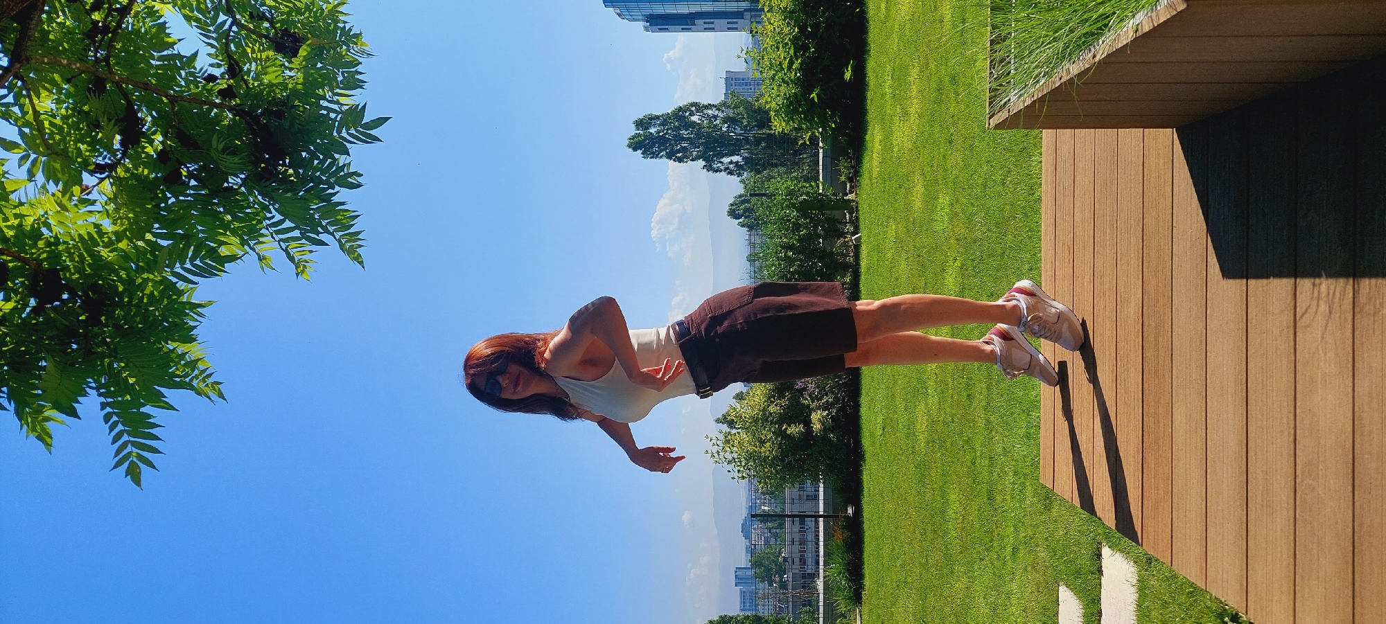 A full-body, side-profile shot captures a woman with auburn hair and sunglasses posing outdoors on a sunny day in Sofia, Bulgaria. She stands on a wooden deck, wearing a white tank top, dark brown shorts, and light-colored sneakers. Her body is angled, with her left arm extended back and her right arm bent, her hands positioned near her waist. The background reveals a well-manicured green lawn, a distant cityscape with buildings and mountains, and lush green trees. The sky is a clear, bright blue with a few scattered clouds. The foreground consists of the wooden deck and a planter box filled with green grass. The image is taken from a low angle, emphasizing the blue sky and the woman's pose.