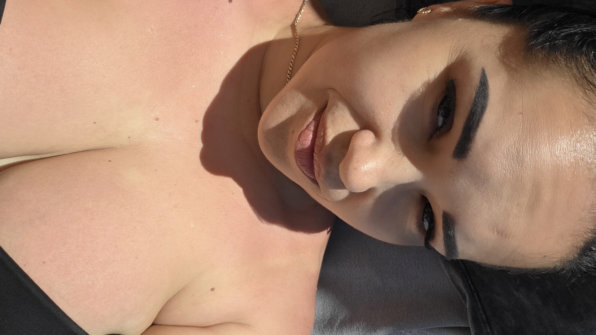 The image is a close-up, outdoor portrait of a woman with dark hair, wearing a gold necklace and a black bikini top. She has dark eyebrows and is looking towards the camera with a slight smile. The lighting suggests it is daytime, with shadows cast across her face and upper body, indicating the sun is high. The background appears to be an outdoor setting, possibly a beach or poolside, with a textured gray surface visible. There are no specific landmarks in the image that would indicate a location within the city of Pivdenne, Ukraine.