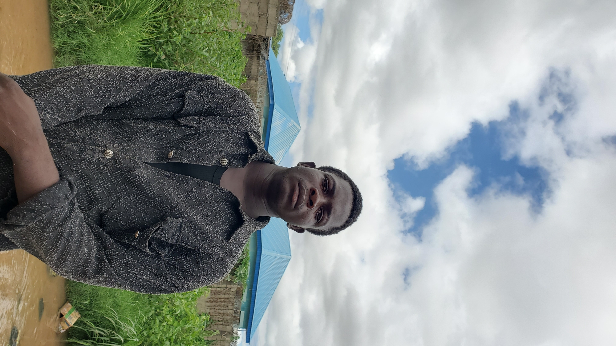 A young Black male stands in front of a cloudy sky with blue patches, his gaze directed forward. He is wearing a dark, textured button-up shirt over a black undershirt. Behind him, two buildings with distinctive blue, pitched roofs are visible, separated by a section of green foliage. A low wall constructed of concrete blocks runs horizontally behind the man and between the buildings. The ground in the foreground appears to be muddy or wet, with some debris visible in the water. The scene suggests an outdoor setting, possibly in a residential or developing area.