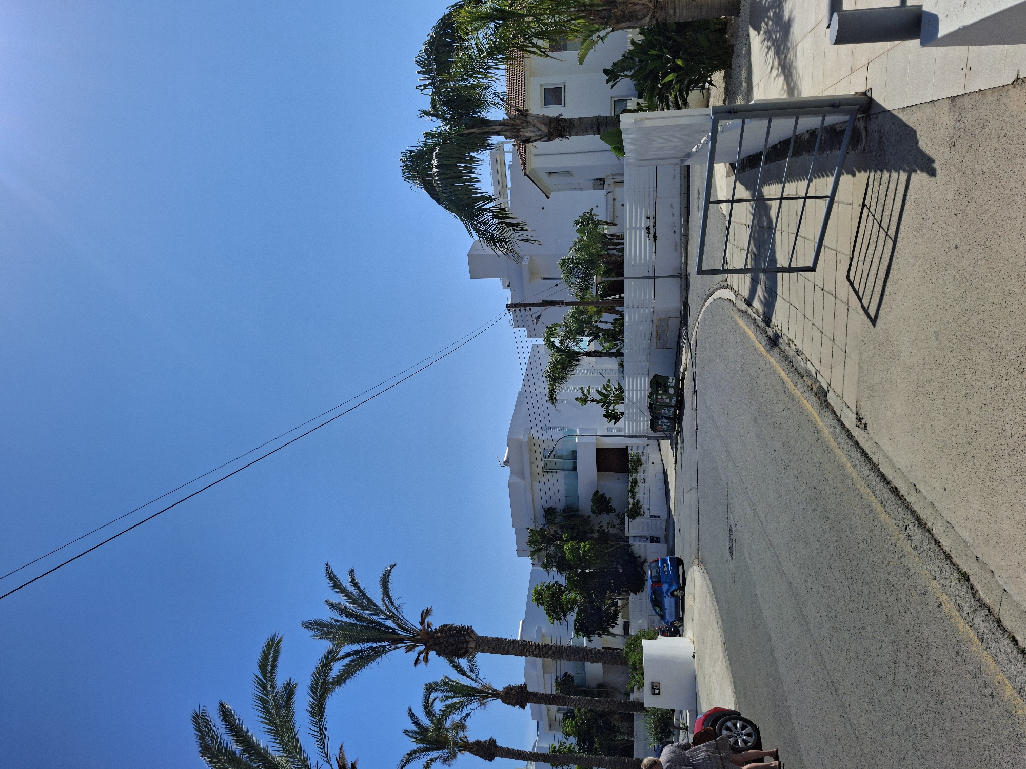The image shows a residential street in Protaras, Cyprus. The street is paved and has a yellow line indicating the road's edge. White buildings with Mediterranean architectural features line the street, interspersed with palm trees and various other vegetation. A blue car is parked along the road. An older woman is positioned in the bottom of the image, presumably on the sidewalk. Overhead, power lines stretch across the clear blue sky. The scene is bathed in bright sunlight, casting shadows along the street and buildings. A metal gate structure is visible near one of the buildings.