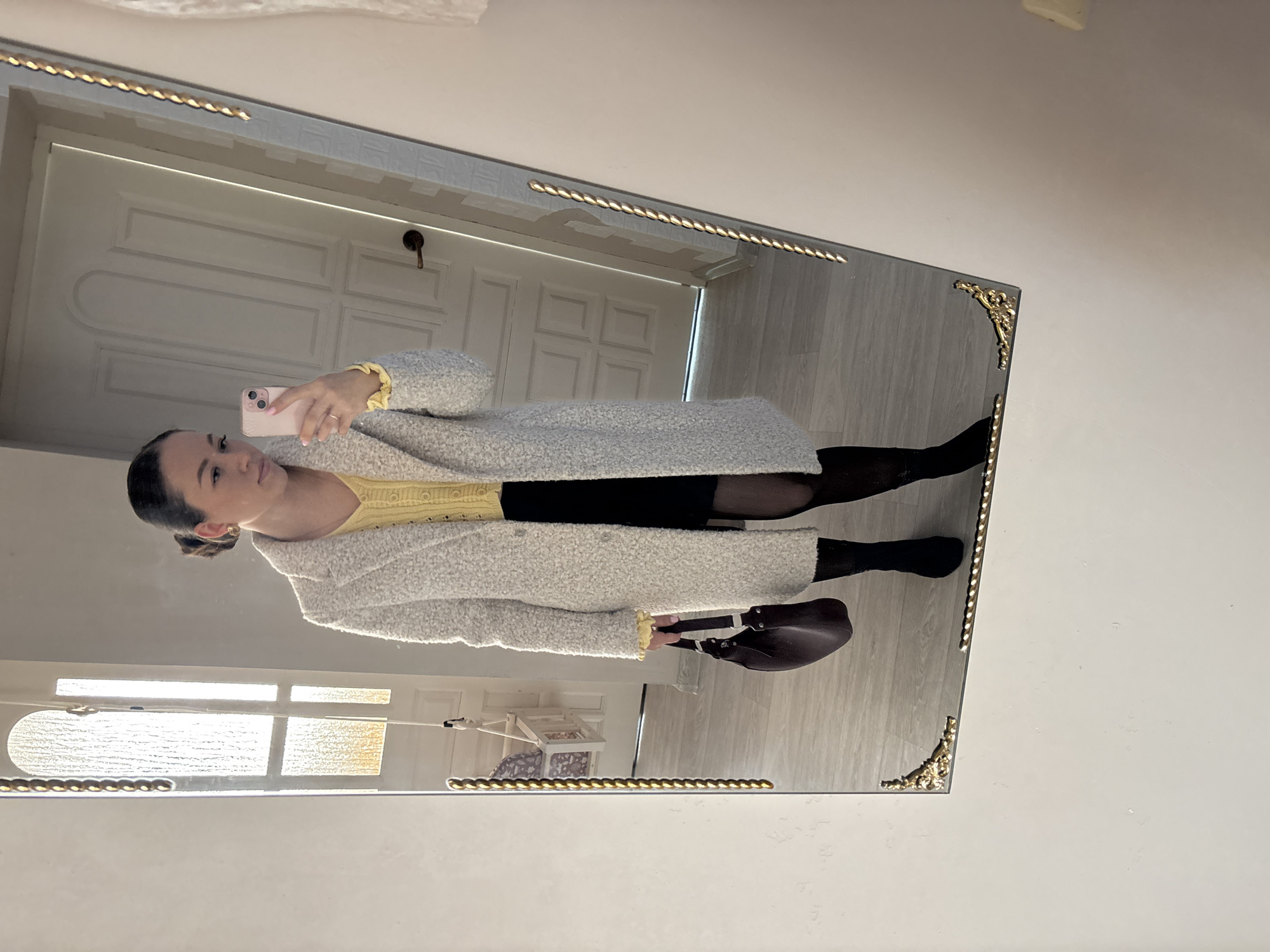 The image is a selfie taken in a mirror. The mirror has a decorative gold frame. A woman is holding her phone up to take the picture. She is wearing a long, white, textured coat, a yellow top, a black skirt, black tights, and black boots. She is also holding a dark brown purse. Behind her in the mirror's reflection is a white door, flooring that appears to be either wood or tile, and a window with a shelf or table underneath. The location is indoors and appears to be a private residence. There is no information available in the image to identify the specific location as being in Ферма, Russia.
