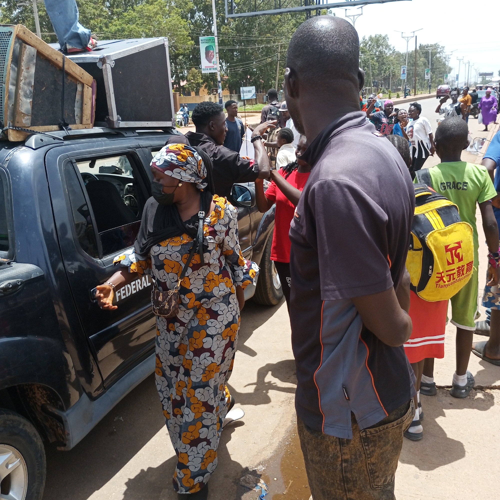 The image appears to depict a scene on a street in Bukuru, Nigeria. A dark-colored pickup truck, marked with the word "FEDERAL" on the door, is parked along the side of the road. The vehicle has large speaker boxes mounted on its roof, and someone is standing on top of them. A woman wearing a colorful patterned dress and a head covering is standing near the truck. She is wearing a face mask and has a purse or bag across her shoulder. A man in a dark shirt and jeans is standing near her, facing away from the camera, with his back to the viewer. Several other people are visible in the background, including a young person with a yellow backpack labeled "Tianyan Education" and a child wearing a green shirt with the word "GRACE". In the distance, there are buildings, trees, and other vehicles, including motorcycles. A sign with a photograph of a person, possibly related to the upcoming election, is visible in the background. The scene suggests a public gathering, possibly related to an election campaign event.