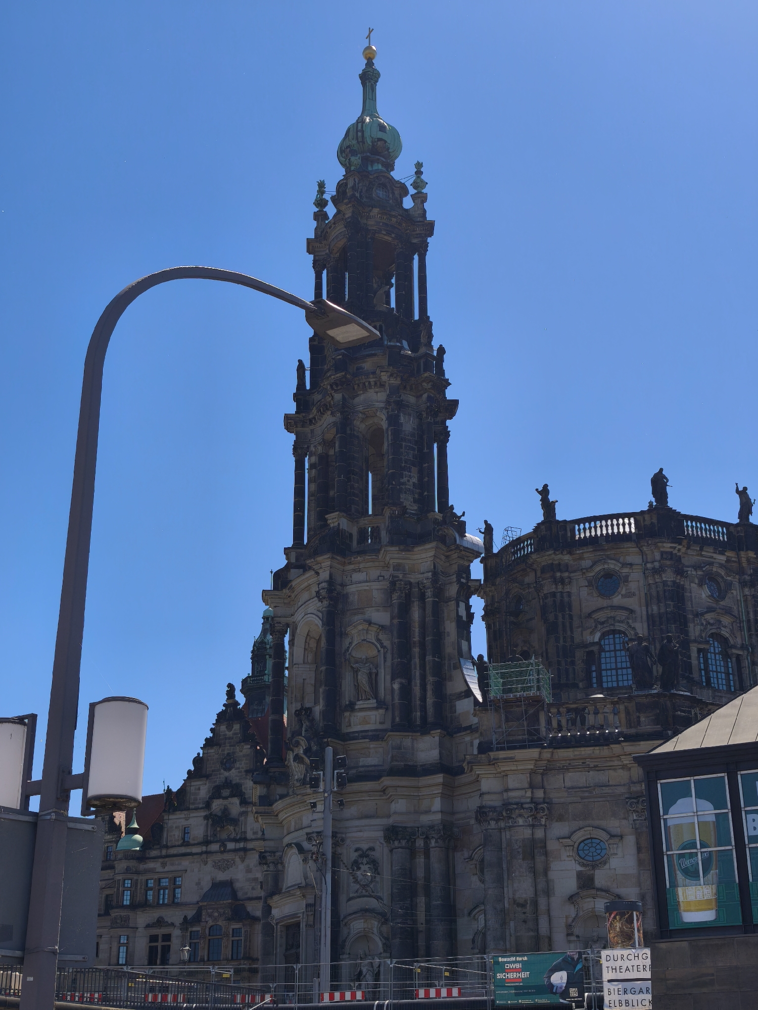 A daytime shot shows the Dresden Frauenkirche from a low angle. The Frauenkirche is constructed from sandstone and has a distinctive dome. Construction or repair scaffolding can be seen on the church. A tall dark gray streetlight with white rectangular lamps is in the left foreground. The sky is a clear blue. Billboards advertising a beer garden are at the bottom right.