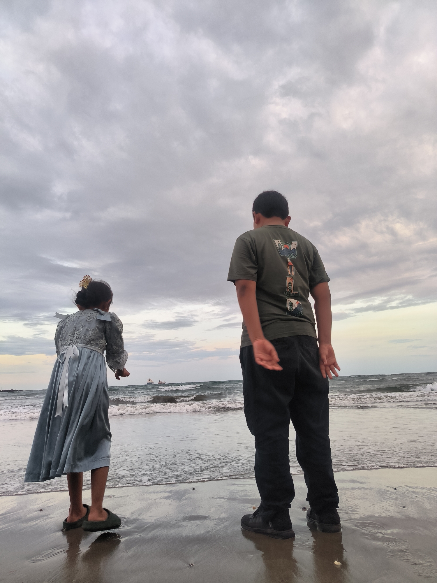 The image captures a back view of two individuals standing on a wet sandy beach at dusk or dawn in Lhokseumawe, Indonesia, looking out at the sea. On the left, a young girl, wearing a light blue-grey dress with a white ribbon tied at the back and dark slippers, stands with her hands slightly open. On the right, an older boy or young man, dressed in a dark green t-shirt with a multi-colored graphic design on the back and dark pants with sneakers, also faces the ocean, his hands hanging loosely. The sky is largely overcast with grey clouds, with a faint lighter, yellowish hue near the horizon on the right. The sea features moderate waves breaking onto the shore, and two distant cargo ships are visible on the horizon line. The wet sand reflects the sky and the figures.