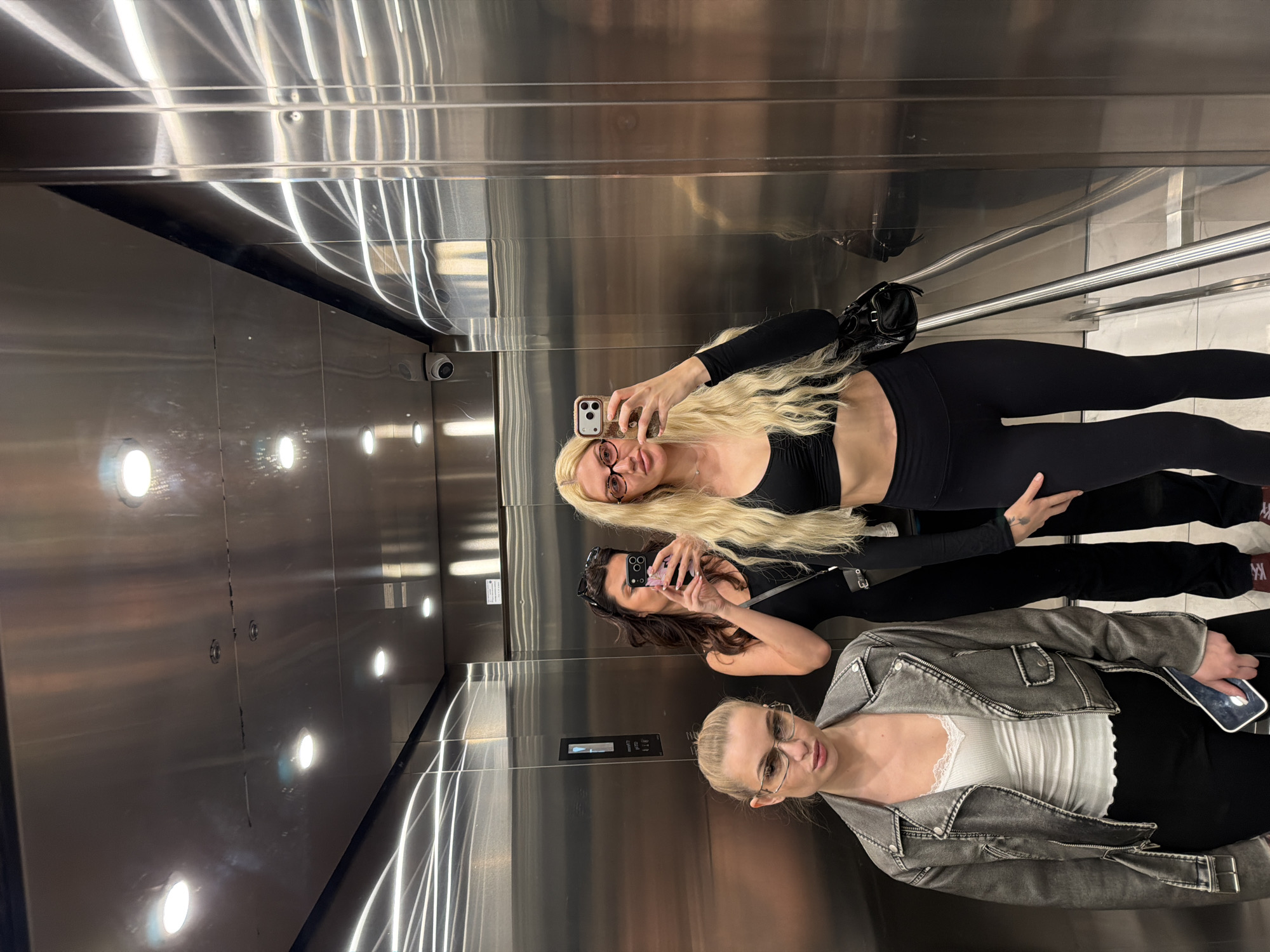 Three women are posing for a selfie in an elevator. The woman closest to the camera is wearing a gray jacket, a white top, and black pants. The woman in the middle is wearing a black top and black pants. The woman farthest from the camera is wearing a black crop top and black leggings. The elevator has stainless steel walls and a white marble floor. A security camera is visible in the upper left corner. A black handbag is worn by the woman in the middle. The elevator appears to be in a modern building, possibly in Belgrade, Serbia, given the provided information.