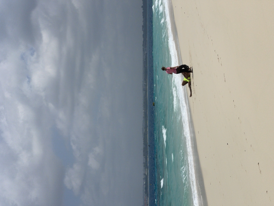 The image depicts a beach in Zanzibar, Tanzania, under an overcast sky dominated by dark grey clouds. The ocean displays turquoise waves breaking onto a light-colored sandy shore, transitioning to a deeper blue further out. A distant landmass or cityscape is visible on the horizon across the water, accompanied by a single yellow buoy. Two individuals are present on the sand: one stands upright wearing a pink shirt and dark trousers, while the other lies near the water's edge in bright neon yellow shorts. The standing person is positioned adjacent to the lower body of the person lying down.