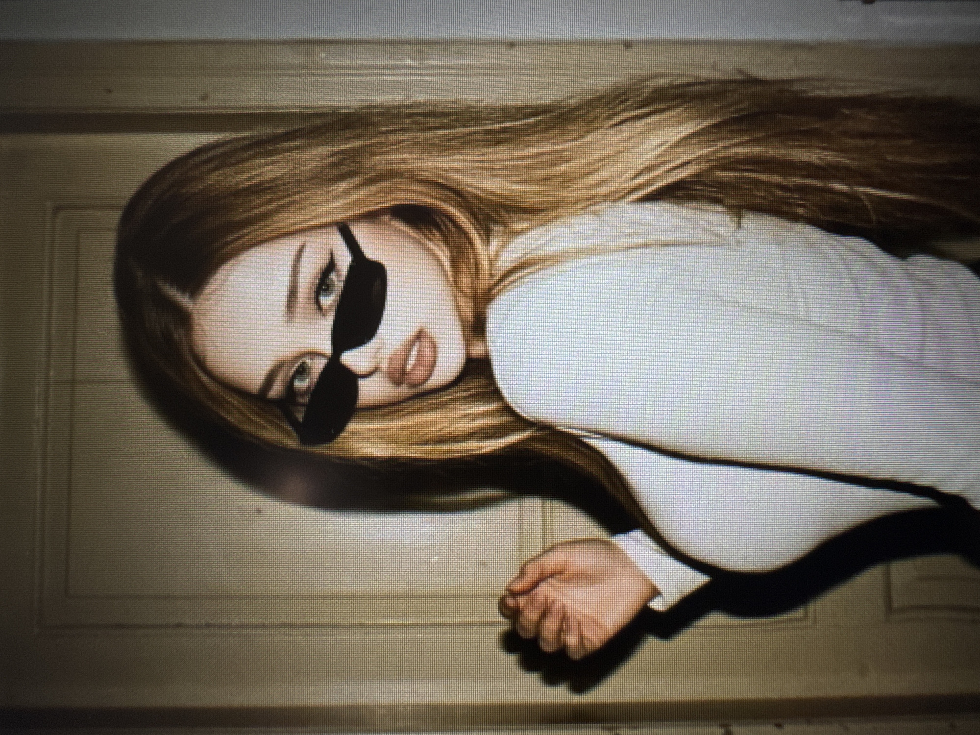 The image is a portrait of a young woman with long, straight, light brown hair, lying down with her head tilted and looking towards the camera. She is wearing a white long-sleeved top and small, black, cat-eye style sunglasses. Her left eye, visible above the sunglasses, appears light-colored, and her lips are slightly parted. Her right hand, partially clenched, is visible in the lower right portion of the frame.
The setting appears to be an indoor space, with the woman positioned against a light brown surface that resembles a wooden door or wall paneling, showing faint vertical lines and texture. The lighting is soft, creating subtle shadows around her hair and arm. The entire image has a noticeable pixel grid pattern, indicating it is a photograph of a digital screen. There is no visible text. The woman's expression is direct and confident, enhanced by the sunglasses.