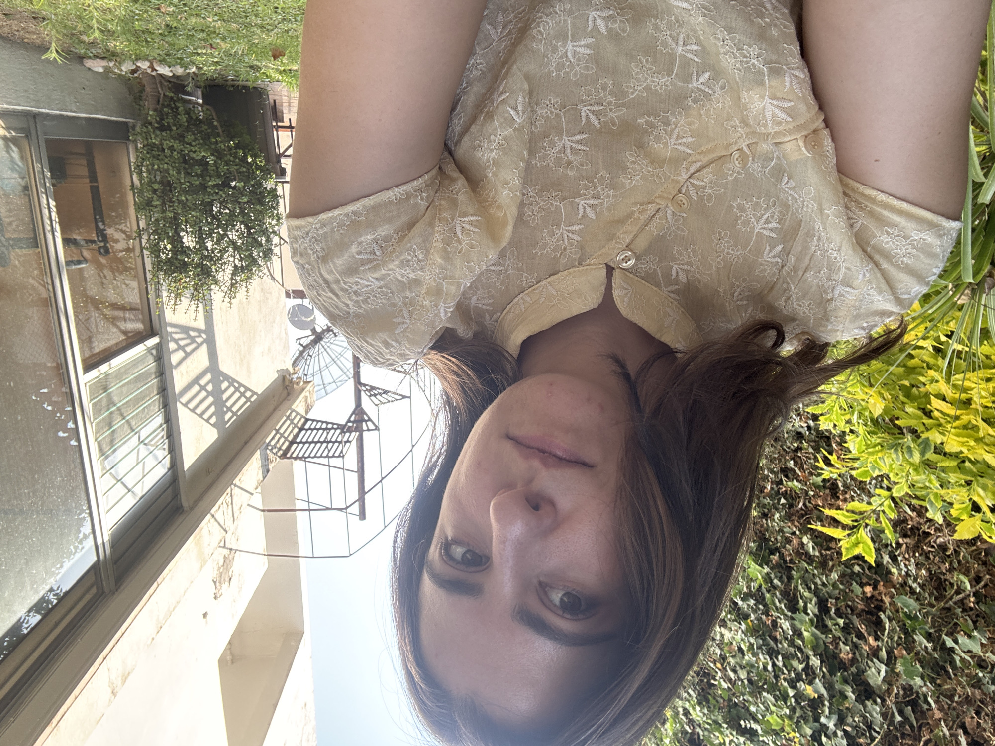 A young woman is positioned facing the camera, wearing a light yellow blouse with floral embroidery and button details at the collar. Her hair is brown and shoulder-length.
The background includes building architecture with a window and security bars. A wrought-iron structure is visible above the building. Green foliage is present, including hanging plants and bushes.
The location is likely within the city of Puebla, Mexico, judging from architectural styles and plant types.