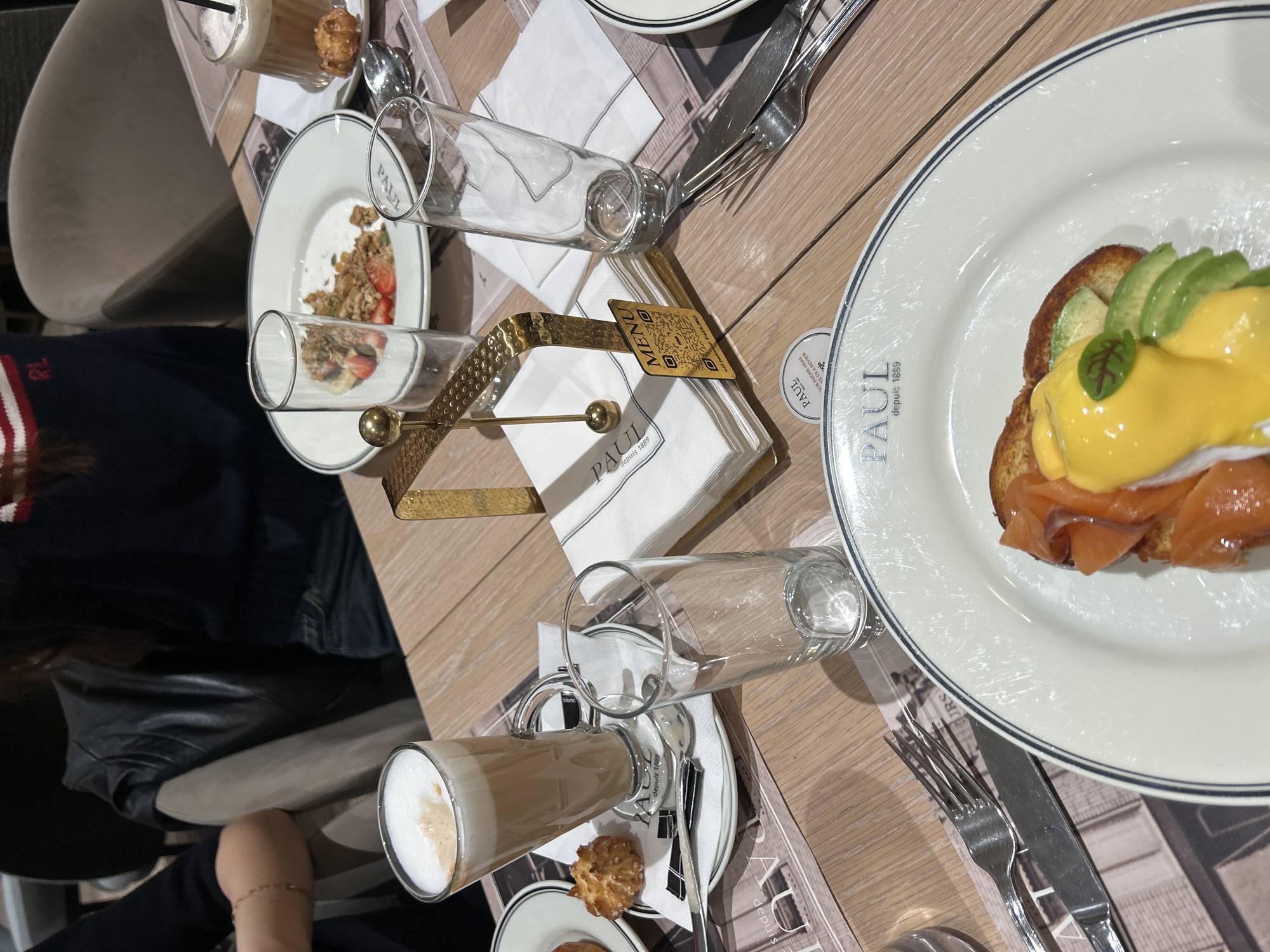 The image presents an overhead view of a table set for a meal, likely breakfast or brunch, at a PAUL cafe or restaurant in Yerevan, Armenia. The wooden table is occupied by several dishes and drinks, suggesting at least two diners.
In the prominent right-hand side, a white plate with a blue rim features Eggs Benedict, prepared with toasted bread, generous layers of smoked salmon, sliced avocado, a poached egg, and a rich yellow hollandaise sauce, garnished with a small green leaf. Adjacent to this, another white plate holds a partially consumed bowl of granola topped with fresh strawberries and what appears to be seeds.
Beverages include a tall glass mug of latte, displaying distinct layers of coffee and frothy milk, served with a small, light-brown cookie on the saucer. Several empty clear water glasses are also scattered across the table. White napkins, cutlery (forks and knives), and white plates, all bearing the "PAUL depuis 1889" branding, are neatly arranged. A central gold-colored, hammered metal napkin holder, also branded "PAUL", holds a small card with a QR code and the text "MENU" and "SCAN YOUR PHONE HERE LEAVE US A REVIEW".
A person's arm, adorned with a delicate gold bracelet, is visible in the lower left portion of the image. Another individual's clothing, featuring a "RL" logo, can be seen in the background. The visible text predominantly highlights "PAUL depuis 1889" across tableware and the napkin holder. Other partially visible text includes "JRS", "depuis", and "blanc". The scene conveys a relaxed, daytime dining experience.