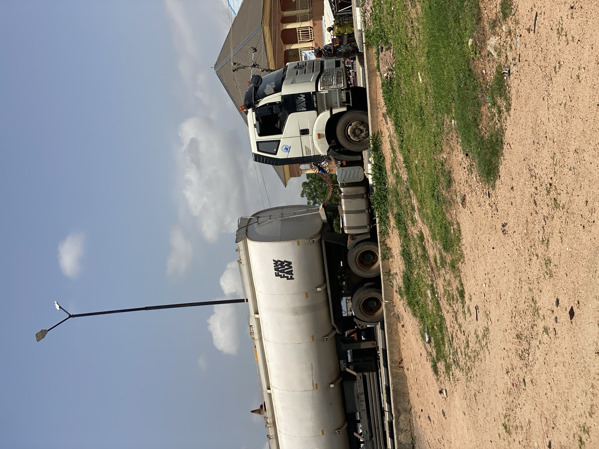 A large white FAW tanker truck is parked on the side of a dirt road in Jalingo, Nigeria. The truck has a single cab with black trim and a large grille. On the side of the tanker are the words "FAW FAW" in large black lettering. The truck is parked next to a patch of green grass and some scattered debris. In the background, there is a building with a light-colored roof and some windows. A street lamp with two lights stands to the left of the truck. The sky is blue with scattered white clouds, suggesting it is daytime. The overall scene depicts a parked truck in a somewhat unkempt outdoor environment.