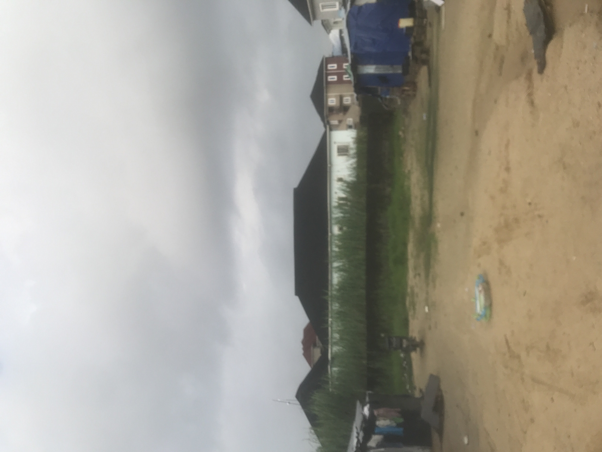 An outdoor, eye-level, medium shot captures a street scene under an overcast sky. The foreground is a sandy, uneven road with a few scattered debris items. In the mid-ground, to the left, is a makeshift structure with clothes hanging on a line outside. Behind this, a thick line of tall green grass separates the foreground from a row of houses in the background. These houses have dark pitched roofs and light-colored walls, with some showing multiple stories. To the right of the grass line, a blue tarp-covered structure, possibly a temporary shelter or stall, is visible. A motorcycle is parked in the middle distance. A colorful inflatable toy lies on the ground in the foreground. The overall atmosphere is somewhat drab due to the cloudy weather.