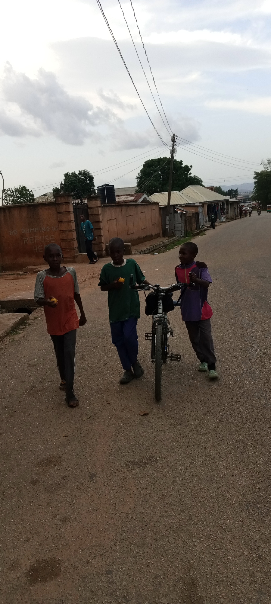 Three boys are on a paved road in Jos, Nigeria. One boy on the left is walking, holding a yellow fruit. The second boy is holding a yellow fruit while standing next to a bicycle. The third boy is leaning against the bicycle while holding a purple object. In the background, there are buildings, power lines, trees, and a mountain range in the distance. A sign on a wall reads "NO DUMPING OF REFUSE HERE". A man stands behind a fence in the background.