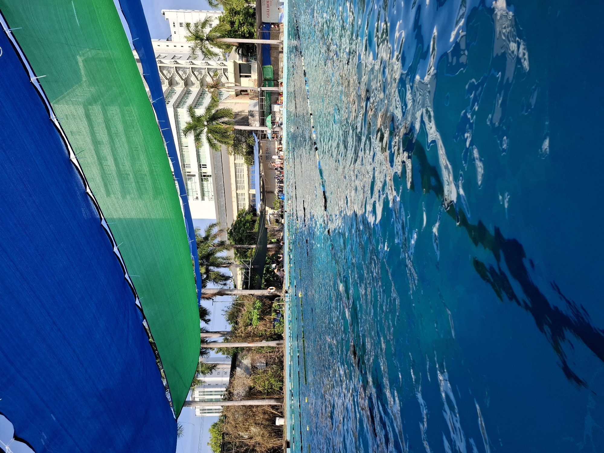 A sunlit outdoor swimming pool is pictured, with lane ropes demarcating swimming lanes. The water is a vibrant blue, with ripples reflecting the overhead shade structure. The shade structure consists of two overlapping layers of fabric, one royal blue and the other green, stretched across the top of the frame. In the background, several palm trees are visible, and beyond them, a modern, multi-story white building with an external staircase. A few people are scattered around the poolside, some sitting and others standing, with a life preserver visible near the edge. The location is identified as a swimming pool in Nha Trang Ward, Vietnam.