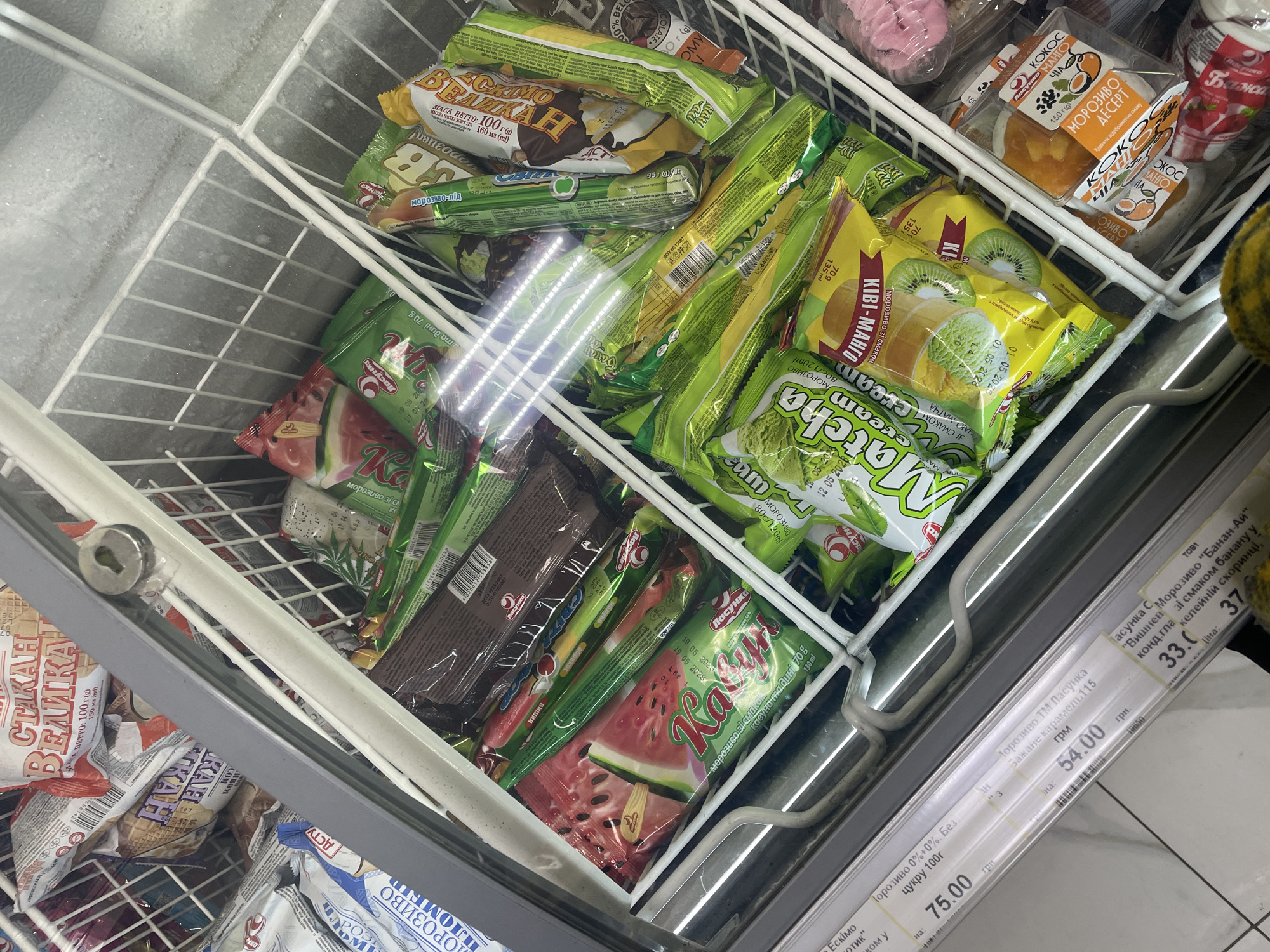 The image captures an overhead view into a grocery store freezer display, filled with various packaged ice cream products. The setting appears to be a supermarket in Rozsoshentsi, Ukraine, indicated by the Ukrainian text on the packaging and price tags.
The freezer is a chest-style unit with a wire rack interior, and some frost is visible on its walls, suggesting cold temperatures. The primary subjects are an assortment of ice cream bars and desserts, predominantly in bright green, yellow, and red packaging, along with some brown and white. The packages are densely packed into compartments within the freezer.
Several types of ice cream are visible, including ice cream on a stick and ice cream desserts. Notable flavors and packaging designs include "КІВІ-МАНГО" (Kiwi-Mango) with illustrations of the fruits, "Matcha cream" in green packaging, and "Кавун та диня" (Watermelon and Melon) featuring a watermelon slice. Other flavors like "Вишня" (Cherry), "Банан-Ай" (Banana-Ai), and "Кокос Манго" (Coconut Mango) are also identifiable. Many products bear the brand name "Ласунка" (Lasunka). Some packages also indicate weight, volume (e.g., "70 g", "100 г", "150 мл"), and a few have production or expiry dates like "01 05 2028" or "19 05 2028".
Price tags are affixed to the side of the freezer, displaying prices in Ukrainian Hryvnia (грн). Visible prices include 54.00 грн for "Морозиво ТМ Ласунка Бажане карамель 115", 75.00 грн for "Морозиво 0%+0%. Без цукру 100г", and 33.00 грн and 37.00 грн for "Ласунка С "Вишнев Морозиво "Банан-Ай". The scene depicts a typical retail environment, showcasing frozen goods for sale.