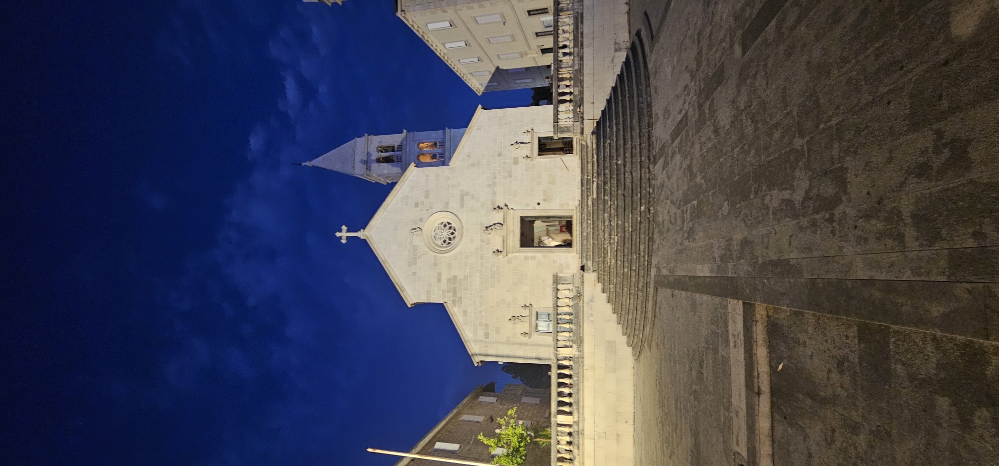 The image captures a nighttime view of a stone church facade with a bell tower. The sky is a deep, dark blue with some cloudy textures visible. The church, constructed from light-colored stone, features a prominent gable with a cross at its apex. A circular stained-glass window is visible above the main entrance. Below this, a rectangular window reveals a glimpse of the interior, with artificial light illuminating the space. To the right of the church, a wide set of stone stairs ascends, leading towards the building. The foreground is dominated by a textured stone surface, likely a plaza or pathway. In the background, parts of other buildings are visible, including a multi-story structure with uniformly spaced windows. A lone, small tree with green foliage is situated to the lower left, adding a touch of nature to the architectural scene. The overall atmosphere is serene and somewhat solemn, enhanced by the dim lighting and the quiet of the night. The scene is set in Grad Supetar, Croatia. There is no visible text in the image.