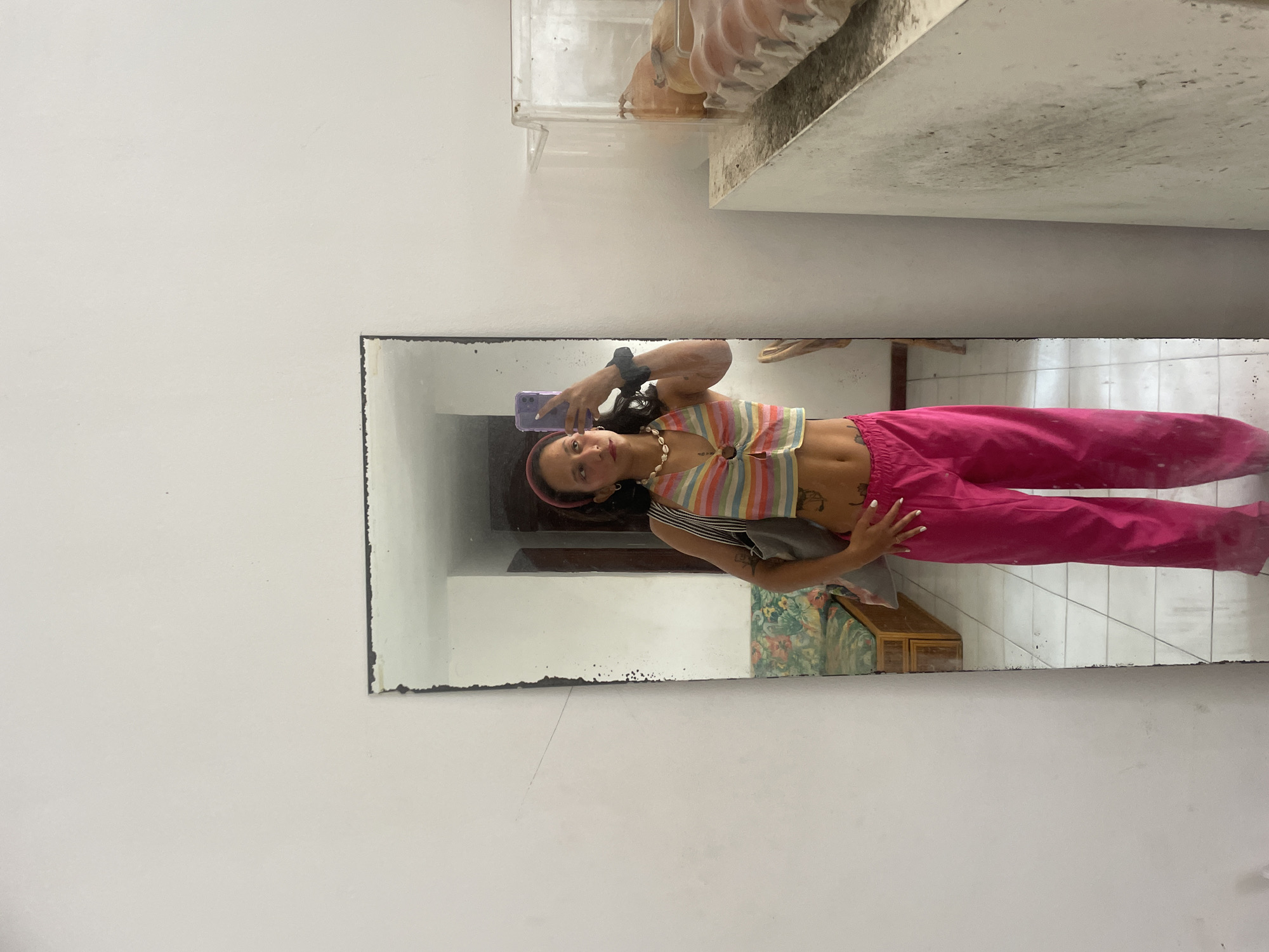A young woman, with dark curly hair and a pink headband, stands in front of a full-length mirror, taking a selfie with her phone. She is wearing a colorful striped crop top and bright pink pants. A grey bag is slung over her left shoulder, and she wears a shell necklace. Her reflection shows her in what appears to be an indoor setting with tiled floors and white walls. To the right of the mirror, a clear plastic container filled with eggs and some squash is visible on a counter. The background reflected in the mirror includes a patterned couch and wooden furniture, suggesting a domestic or somewhat casual environment. The overall scene is a snapshot of a person documenting their outfit in a residential or studio space within the city of Porlamar, Venezuela.