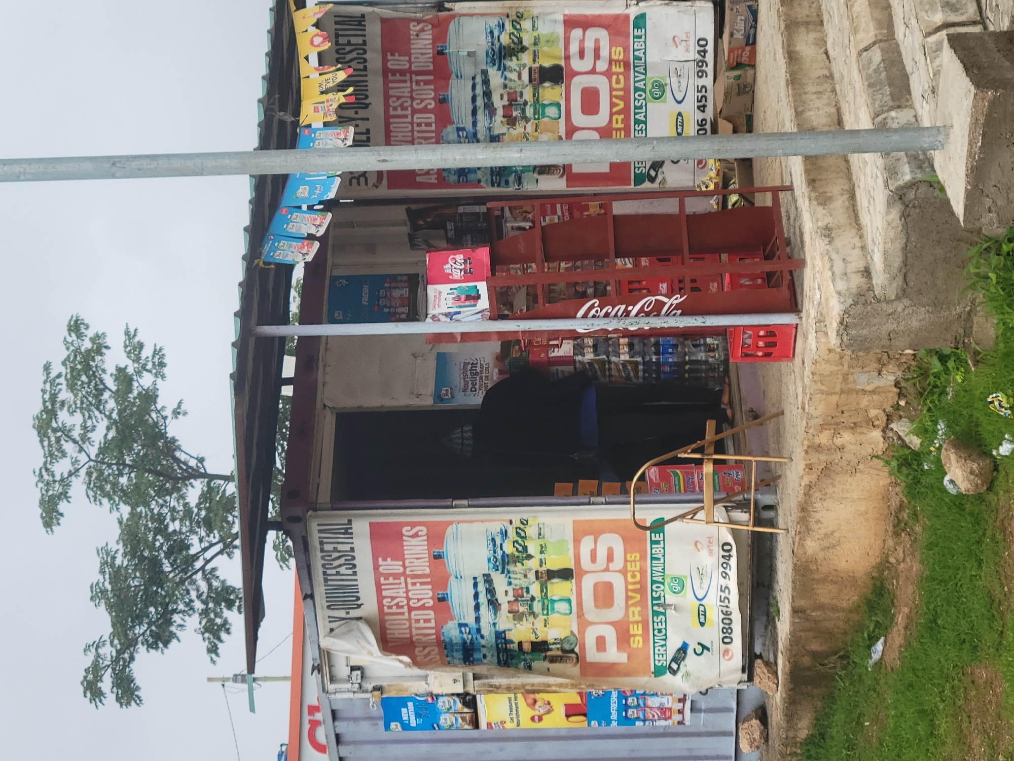 A busy roadside stall, possibly a container shop, is filled with various products, including soft drinks and bottled water. A large poster advertises "Wholesale of Assorted Soft Drinks" and "POS Services," with a phone number "08061559940" and logos for Glo, MTN, and Airtel. A person is partially visible inside the stall, with their back to the camera. In front of the stall, a red Coca-Cola display rack stands, along with a gold folding chair. The ground is rough and uneven, with patches of grass and scattered rocks. The overall scene suggests a commercial hub in an urban setting.