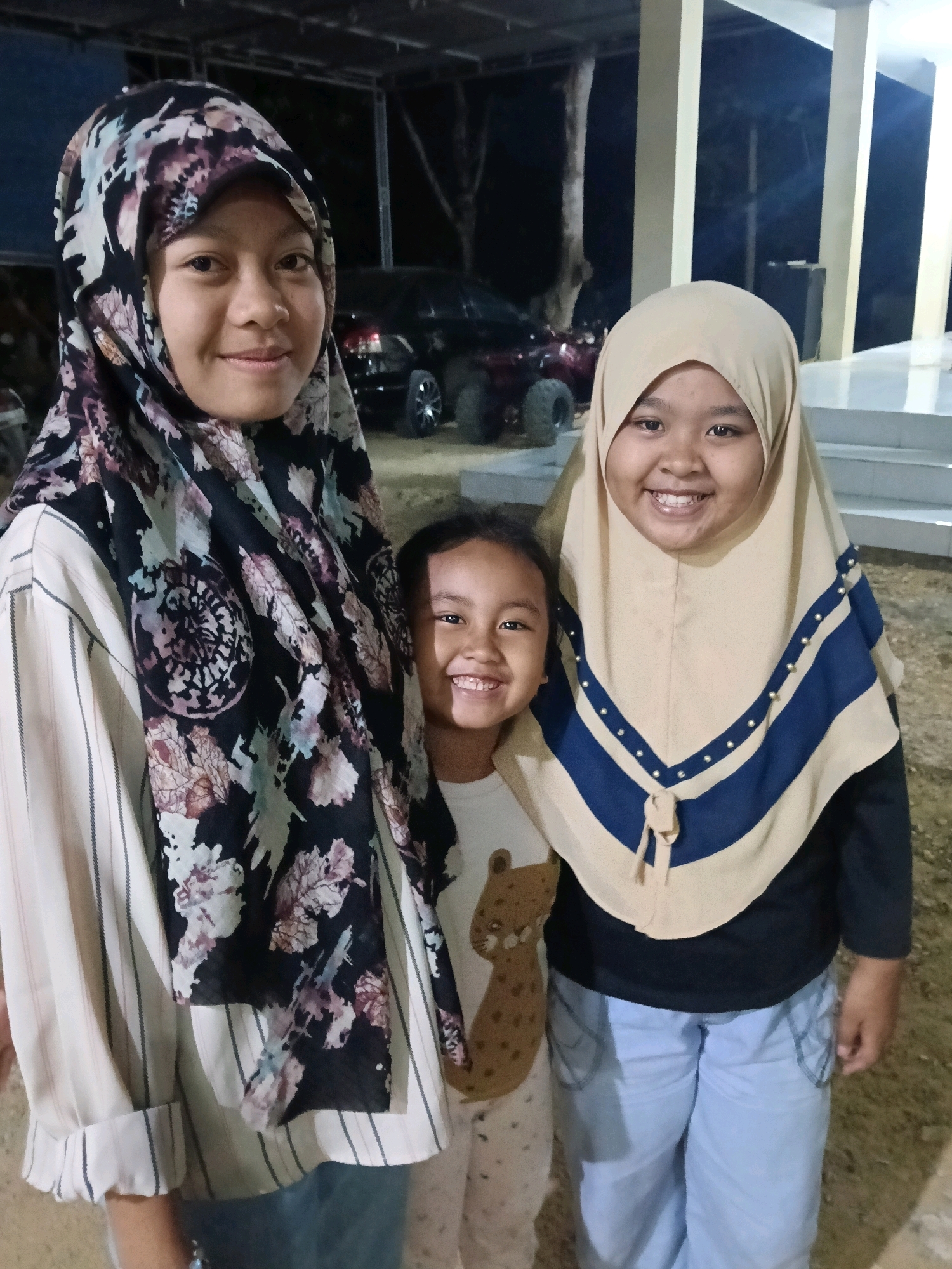 Three individuals, two adult females and one young female, pose for a photograph outdoors at night. The central adult female on the left is wearing a black and floral-patterned hijab and a striped long-sleeved shirt. To her right, a younger girl is visible from the chest up, wearing a white shirt with a brown animal graphic. The second adult female on the far right is wearing a light beige hijab with blue and gold accents and a dark long-sleeved shirt, paired with light blue pants. The background suggests an outdoor setting with structures that appear to be part of a covered area with pillars, possibly a building entrance or shelter, and some vehicles are visible in the distance, including a red ATV. The ground is covered in dirt or gravel. The scene is lit by artificial lighting, creating a nighttime atmosphere. The location is identified as Bulangan Barat, Indonesia.