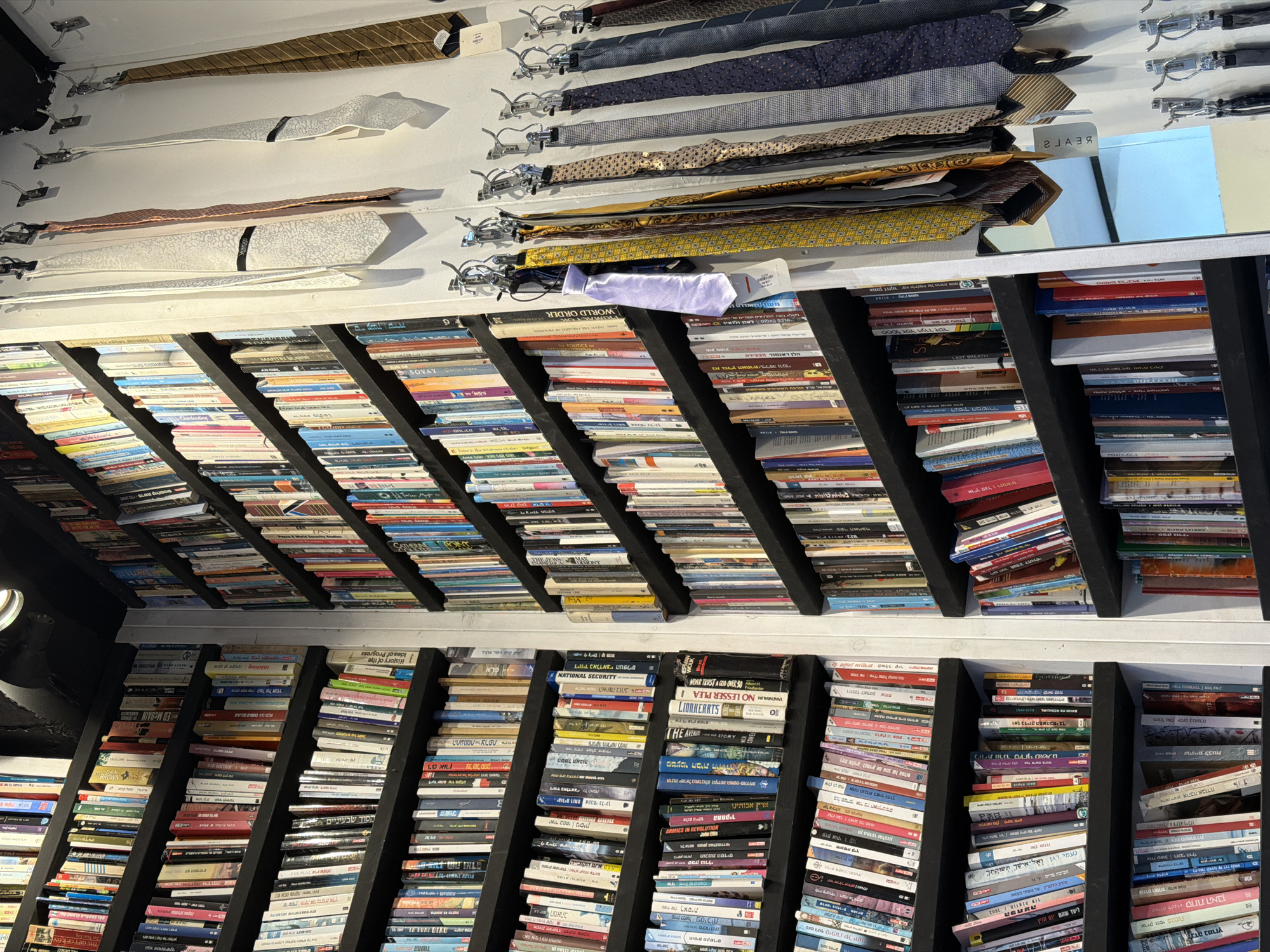 This image captures a unique interior display, likely within a boutique or bookstore in Tel-Aviv, Israel. Numerous neckties and bow ties are neatly hung from hooks, suspended from what appears to be the ceiling or an overhead structure. These ties exhibit a variety of patterns, including stripes, dots, and solid colors like gold, brown, blue, and purple. One purple bow tie clearly displays a "REAL" tag.
Below the hanging ties, an extensive collection of books is densely arranged on black shelving units. These shelves are angled, suggesting they might be part of an unusual ceiling-mounted bookshelf system or a distinct architectural feature. The books are packed closely, with many spines visible, indicating a large inventory or collection.
The visible text on the book spines is a mix of Hebrew and English titles, including "WORLD ORDER," "NATIONAL SECURITY," "LIONKERRTS," "ARMIES IN REVOLUTION John Ellis," "Idea of Progress," "ED MCBAIN THE GIPF," "LAST BREATH," and various Hebrew titles such as "הנסיך המשורר" (The Poet Prince), "בשורות טובות" (Good News), "אהבה אבודה" (Lost Love), "האישה" (The Woman), "התחקיר" (The Investigation), and "תרבות ישראל במערבולת הקידמה הפוסט-מודרנית" (Israeli Culture in the Whirlwind of Post-Modern Progress). A subtle reflection of "REAL" (or part of it) is visible on a shiny surface above some books, aligning with the "REAL" tag on the bow tie.
The overall scene presents an eclectic and creatively arranged space, combining fashion accessories with literature, suggesting a curated retail environment or a highly personalized collection. The shot is taken from a low angle, looking up towards the ceiling, emphasizing the abundance and unique display of both items.