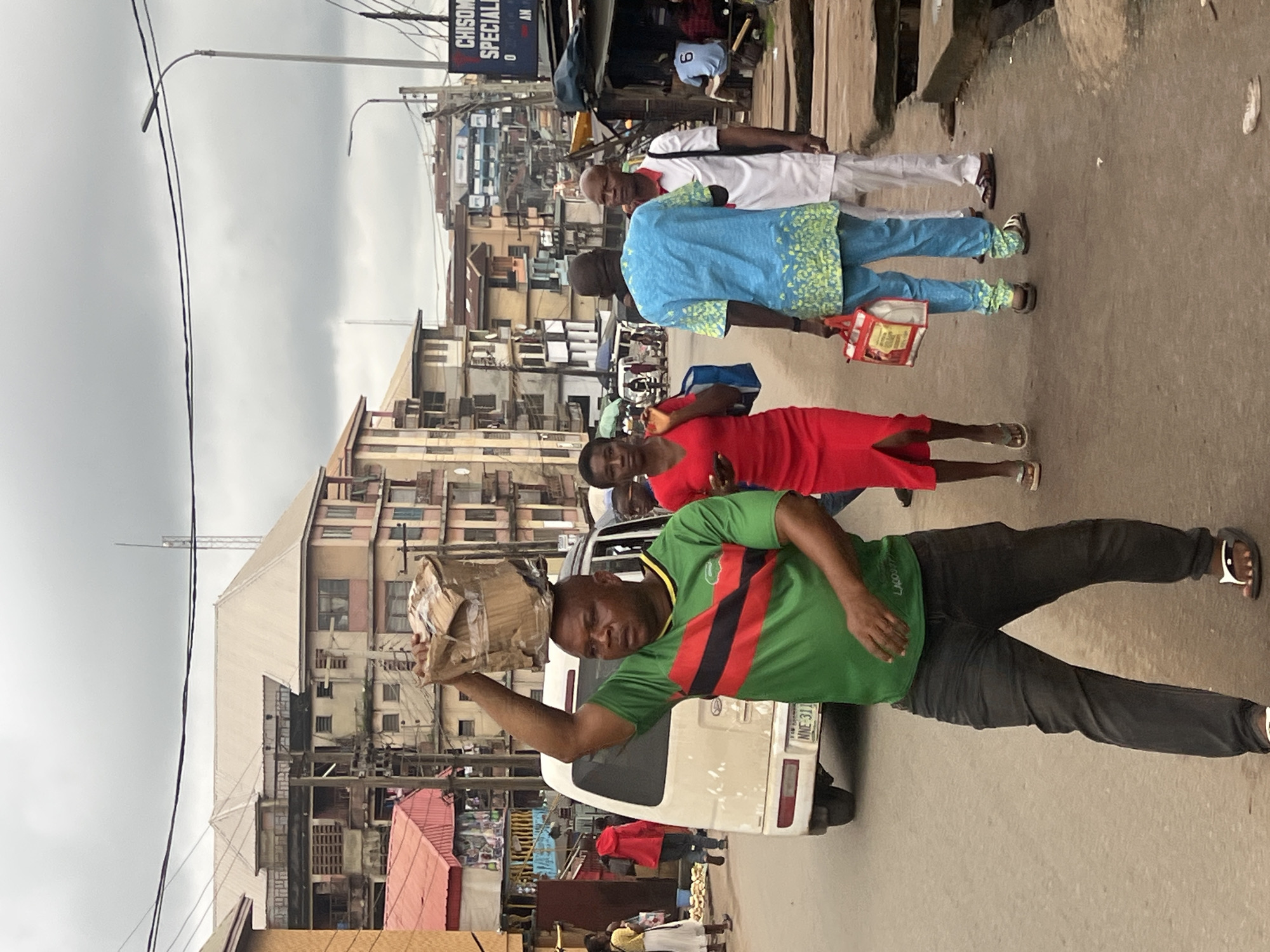 The image depicts a street scene in Nkpor, Nigeria. In the foreground, a man wearing a green and red striped t-shirt and dark jeans carries a large cardboard box on his head. He is walking on a paved road. Behind him, a woman in a bright red dress walks away from the camera, holding a blue bag. To the right of the man, a person in a blue patterned shirt and blue pants walks towards the background, carrying a red and white bag. Another person in a white outfit walks beside them. The background shows multi-story buildings, some with balconies, and a van is parked on the left side of the road. Several utility poles with wires are visible, and the sky is overcast. The overall environment appears to be an urban commercial or residential area.
