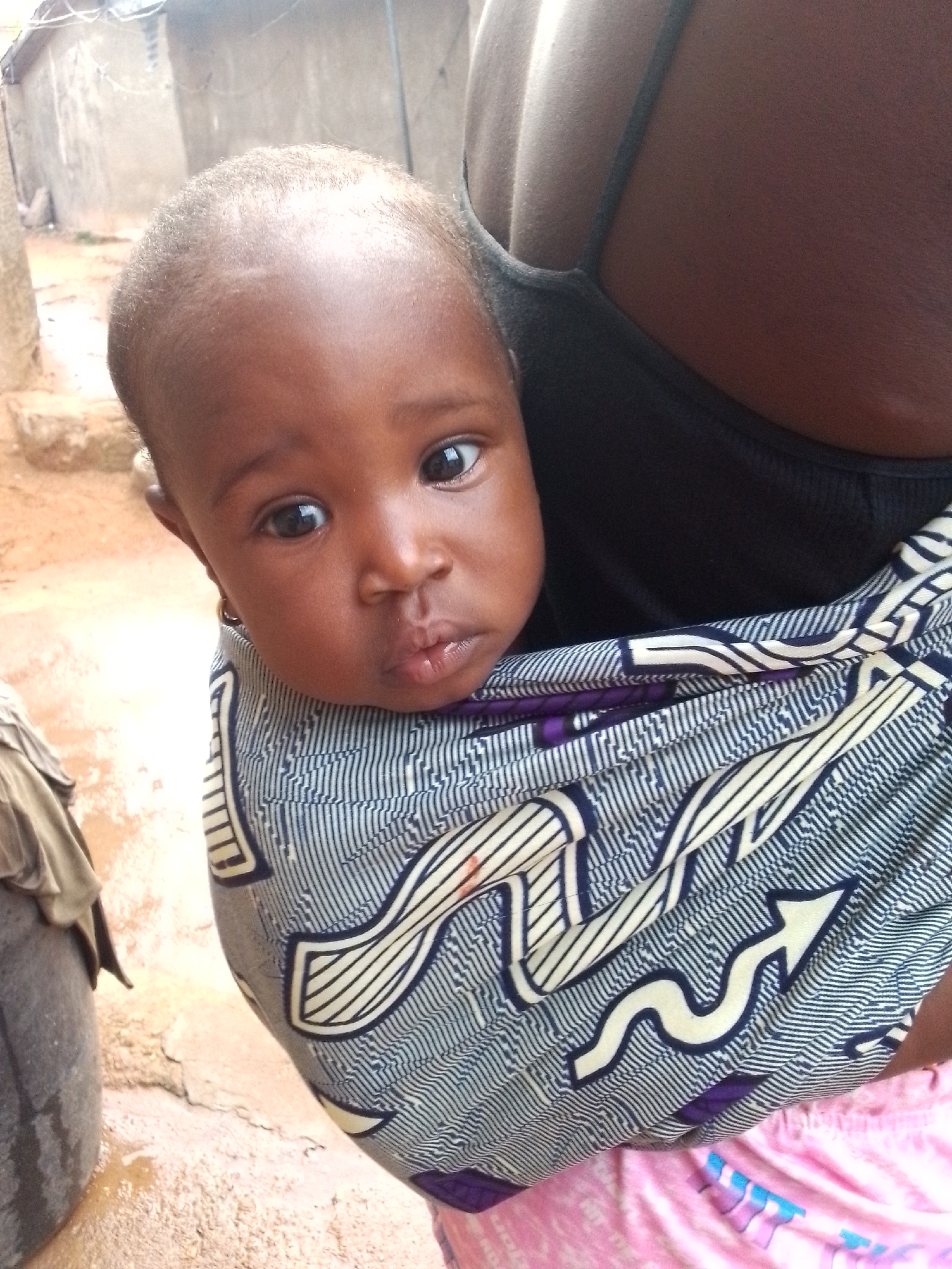 A baby with dark skin and a small gold earring is carried in a sling against the chest of an adult. The baby is looking directly at the camera. The sling is blue and white with an arrow-like design. The adult wears a black tank top and pink patterned shorts. The location appears to be outdoors in Narayi, Nigeria, with concrete buildings in the blurred background. A gray barrel is visible on the left. The ground is light brown.