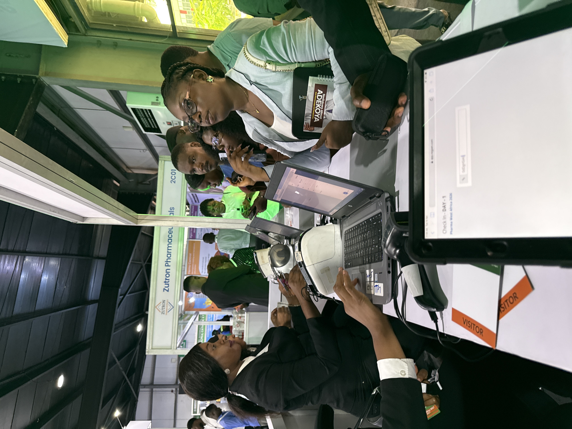 The image shows a group of people at what appears to be a trade show or conference for pharmaceuticals, likely in Itirin, Nigeria, as indicated by "Pharma West Africa 2026". A woman in a black suit is operating a laptop and a barcode scanner, seemingly checking in visitors. A tablet in the foreground displays a check-in screen. Another woman wearing glasses and holding a badge with the name "Similoluwa Adekoya" is interacting with the check-in process, as are other people standing behind her. Signage for "Zutron Pharmaceuticals" is visible in the background. The scene suggests an active check-in point for visitors at the event. "Visitor" is labeled on signs on the table.