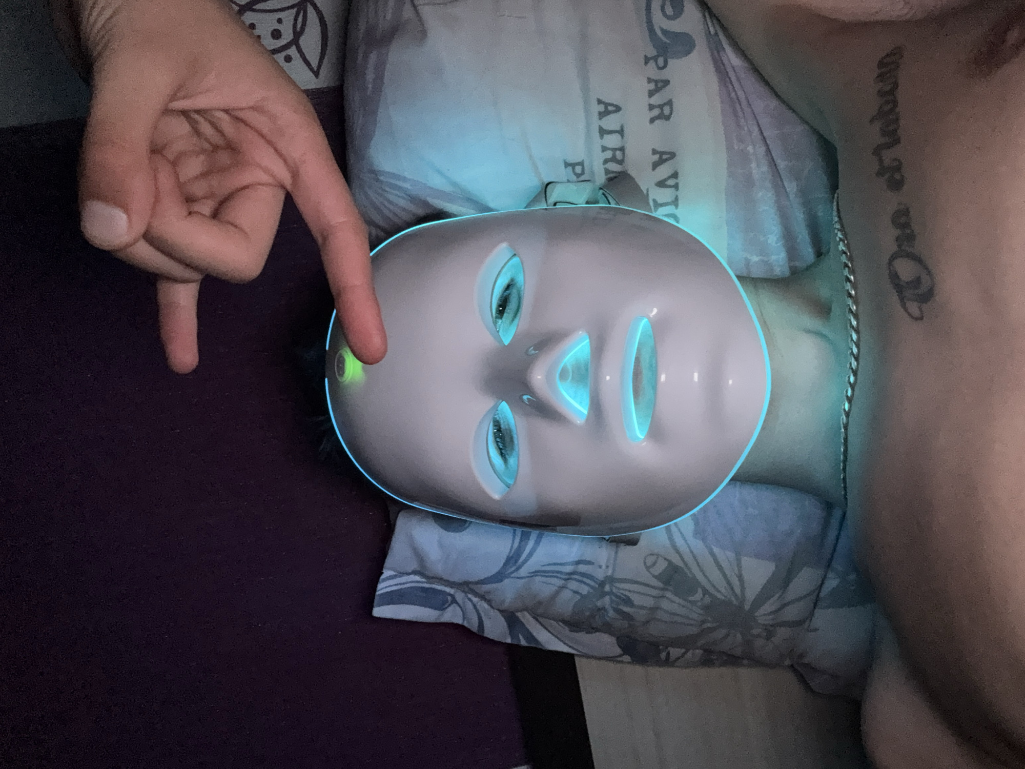 A person lies on their back wearing a white LED facial mask with blue and green lights. A hand with two fingers extended points towards the top of the mask. The person has a tattoo on their chest and is wearing a silver chain necklace. The background consists of a purple textured surface and a pillow with a gray floral pattern. The scene appears to be indoors, possibly in a bedroom. No specific location in Stara Zagora, Bulgaria, can be determined from the image.